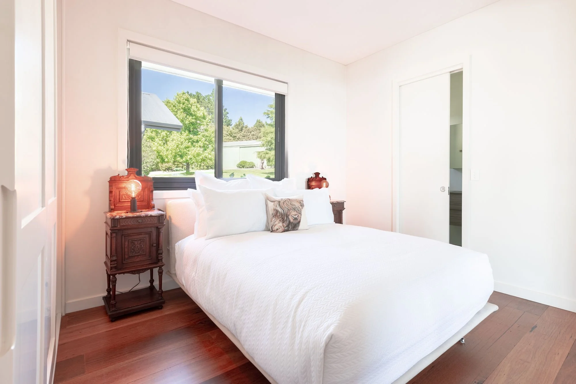 A bedroom with a white bed, wooden nightstands with lamps, a window showing trees and a neighboring house, and a doorway leading to another room.