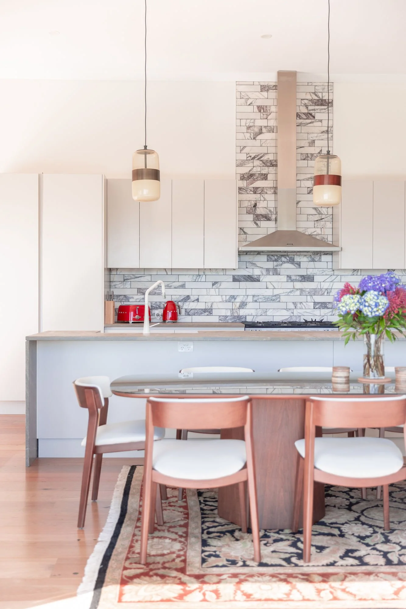 Modern kitchen with white cabinets, marble backsplash, stainless steel range hood, and two pendant lights hanging over the counter. There are red appliances on the counter and a dining area with a table, chairs, and a floral centerpiece.