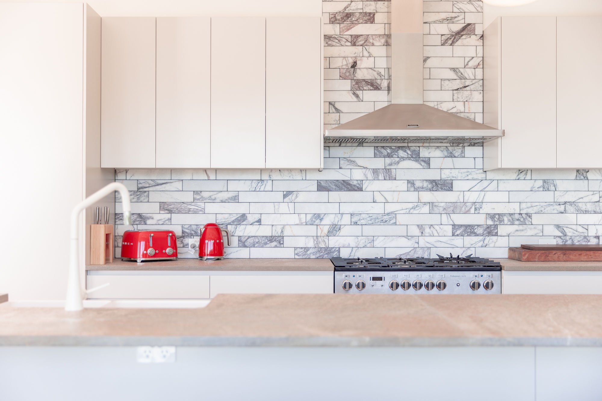 Modern kitchen with white cabinets, a marble tile backsplash, a stainless steel range hood, a stove, a red toaster, and a red kettle on the countertop.