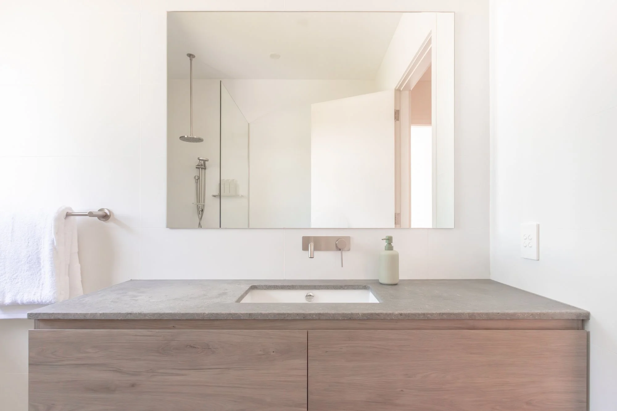 A modern bathroom vanity with a granite countertop, a sink, a liquid soap dispenser, a large mirror, a shower area with a rain shower head and a handheld shower, a towel on a bar, and a door leading to another room.
