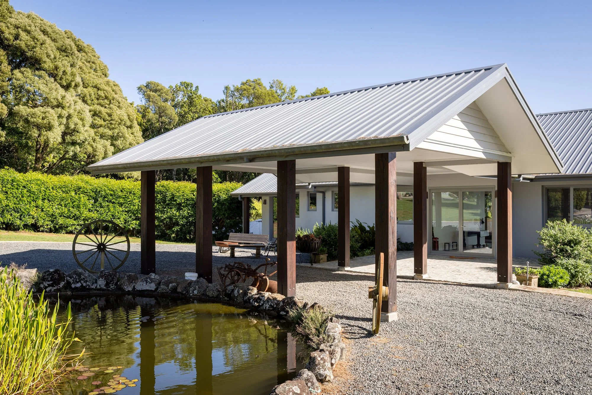 A house with a metal roof, surrounded by greenery and trees, next to a pond with water lilies, and outdoor seating with a bench and rustic wagon wheel decorations.