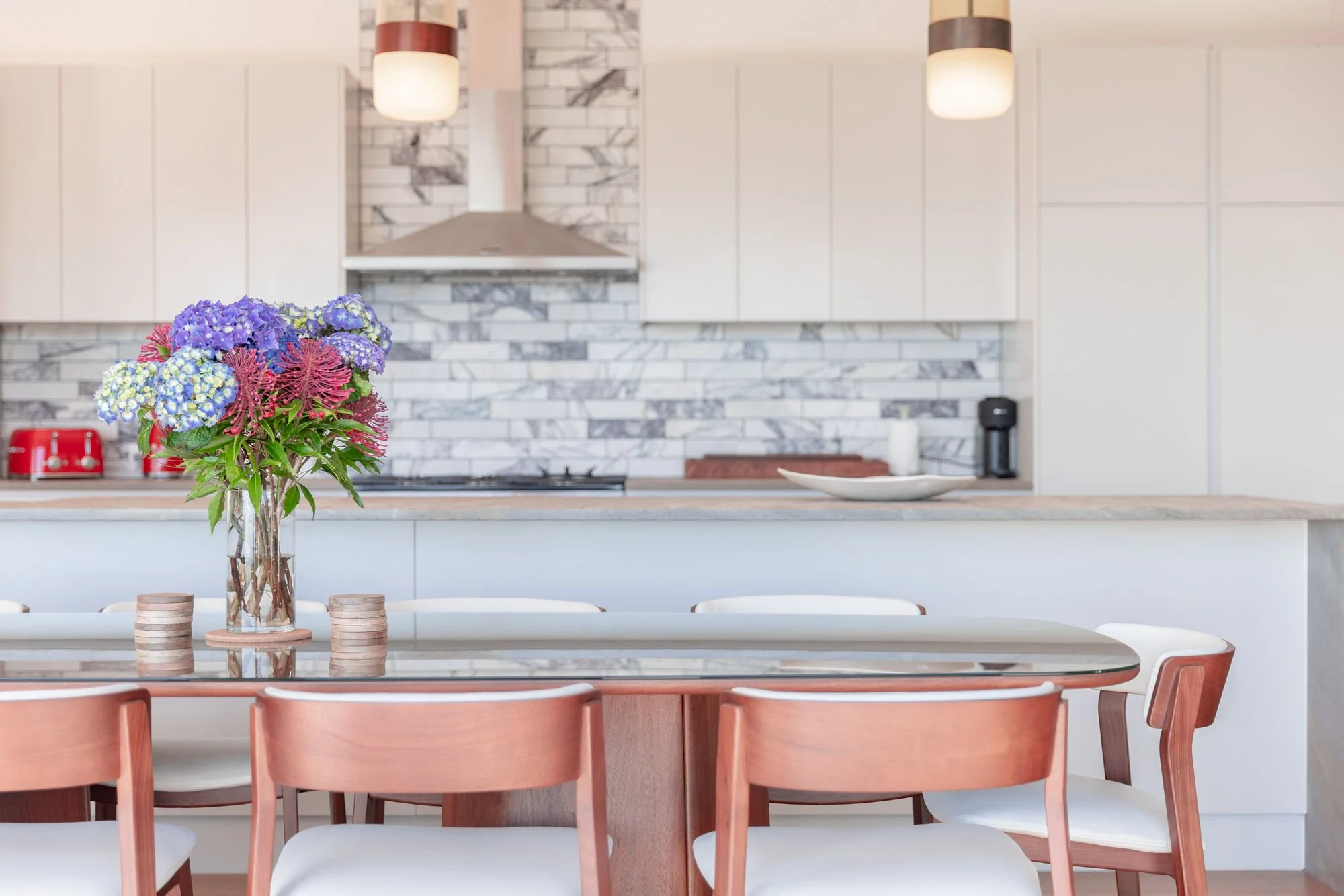 A modern kitchen with white cabinets, marble backsplash, and two pendant lights. A vase of purple and pink flowers sits on the dining table in front.