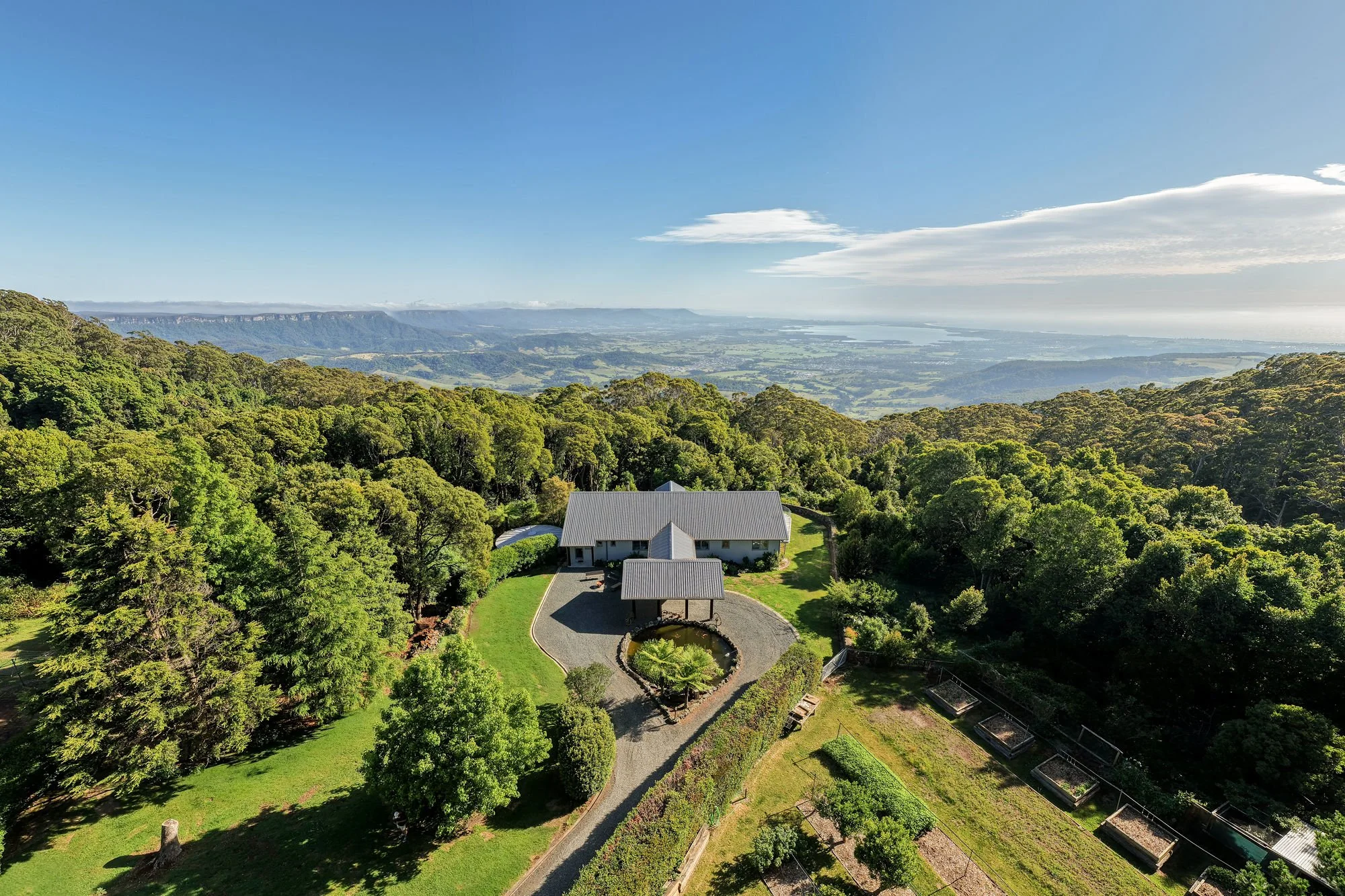 A scenic view of a house surrounded by lush green trees, a garden with a round pond and sunlit landscape, with forest-covered hills and a distant body of water under a blue sky with scattered clouds.