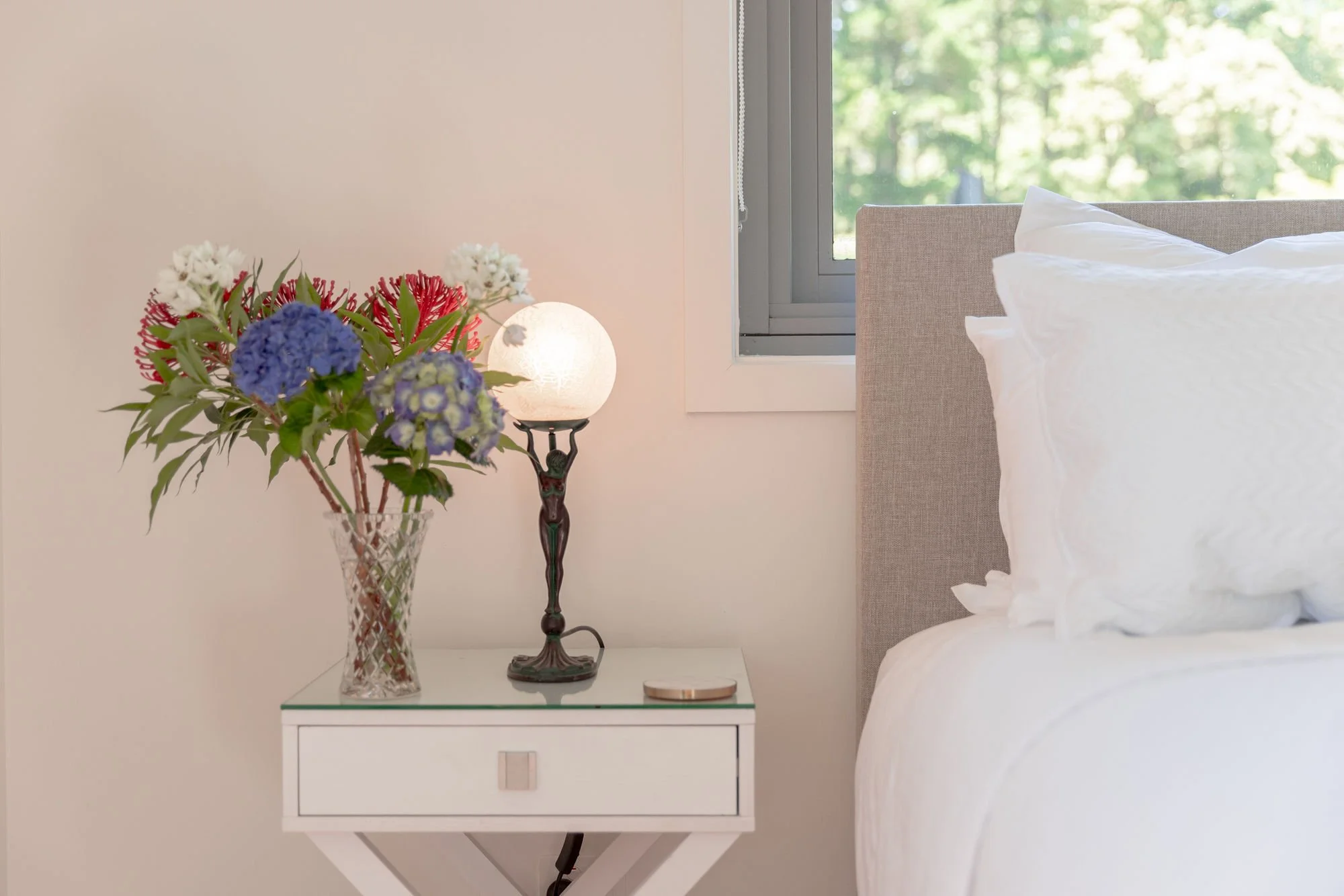 A white nightstand with a glass top holding a floral arrangement in a glass vase, a decorative lamp with a spherical soft white shade, and a small round item. To the right, there's part of a bed with white bedding and a light-colored upholstered head
