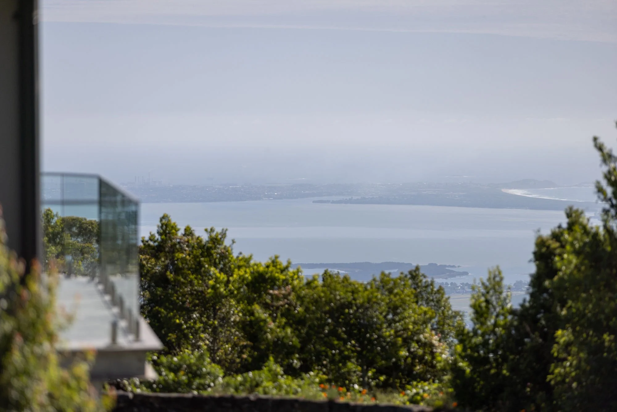 A scenic view overlooking a large body of water with distant land and hills, partially obscured by trees and part of a building with a glass balcony in the foreground.