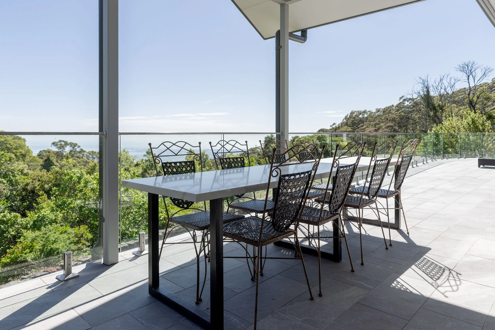 Outdoor balcony with a white table and black metal chairs overlooking a green forest with hills in the distance under a clear blue sky.
