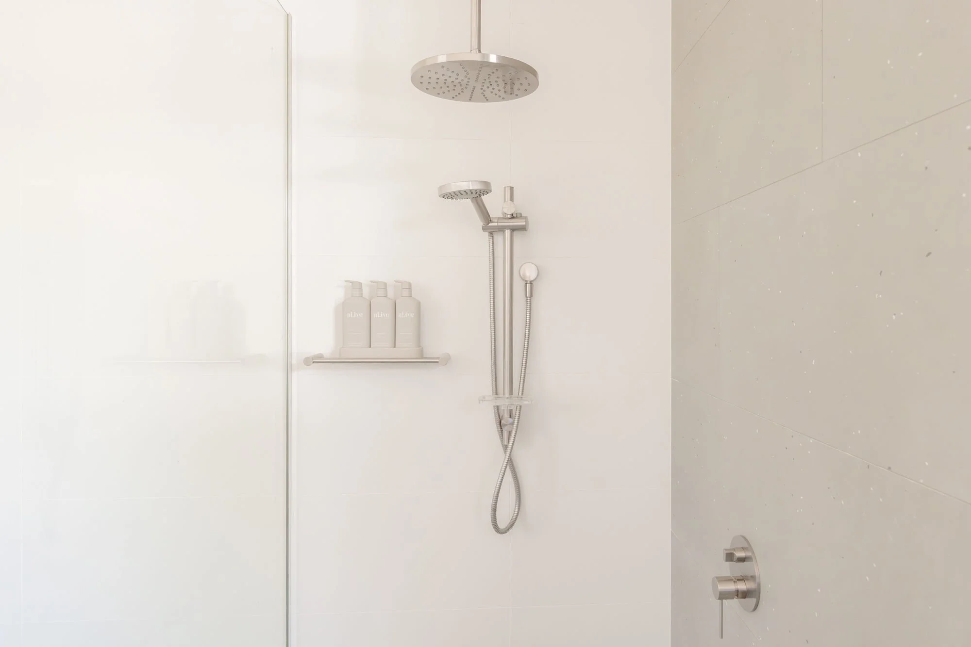 A minimalist shower with white tiled walls, a rain showerhead, a handheld showerhead, a shelf with three bottles, and a control panel on the wall.