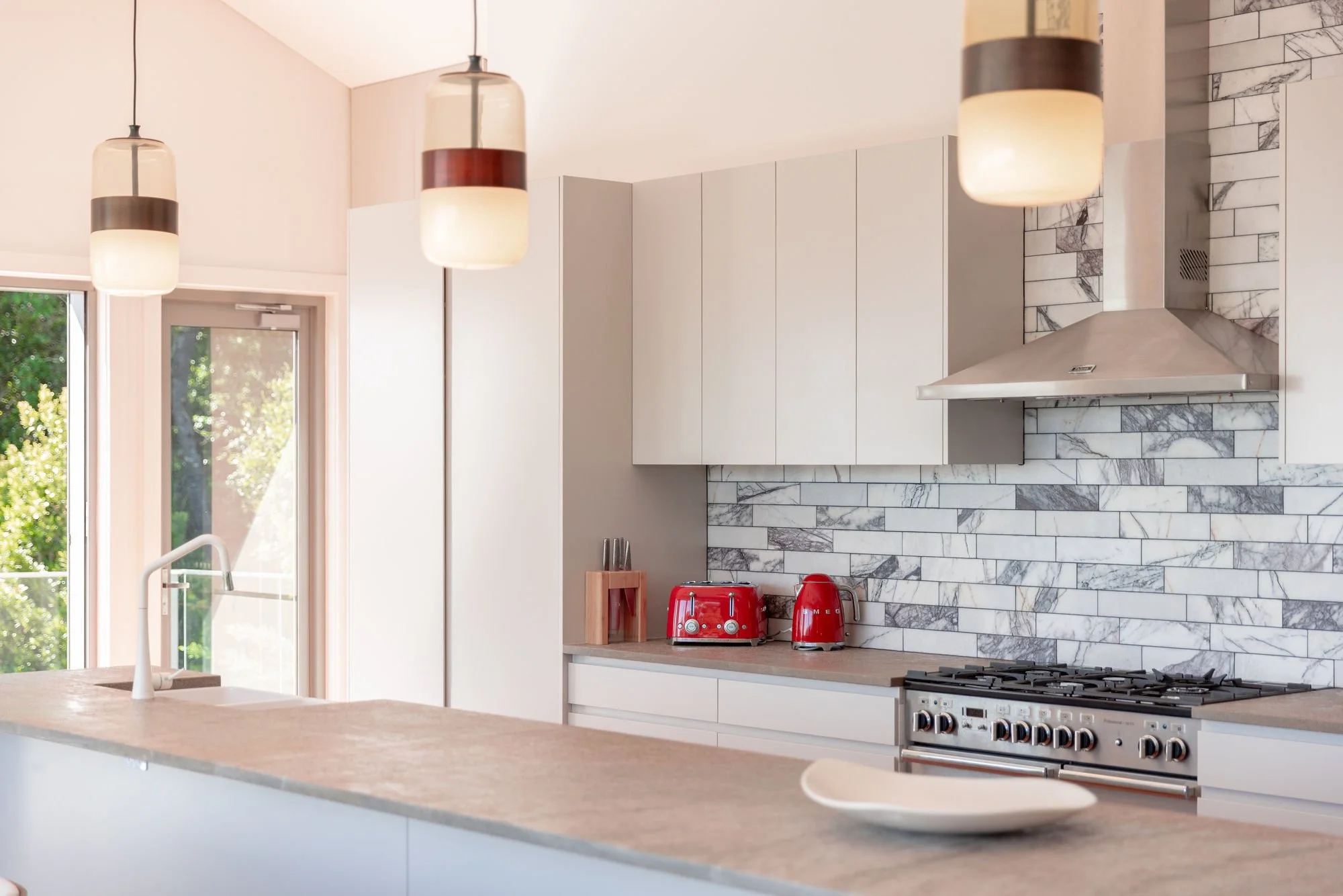 Modern kitchen with white cabinetry, marble backsplash, stainless steel range hood, red kitchen appliances, and pendant lights.