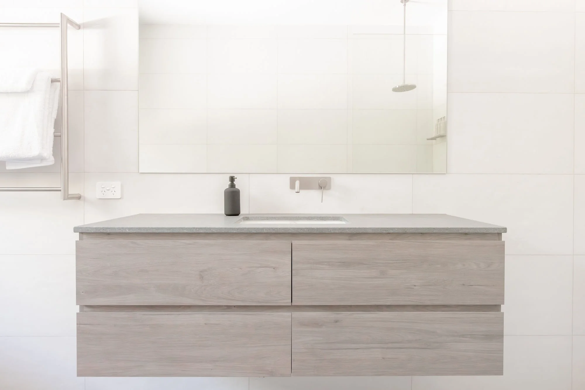 A minimalist bathroom with a floating vanity, large mirror, black soap dispenser, wall-mounted faucet, white tiled walls, a white shelf with towels, and a showerhead.