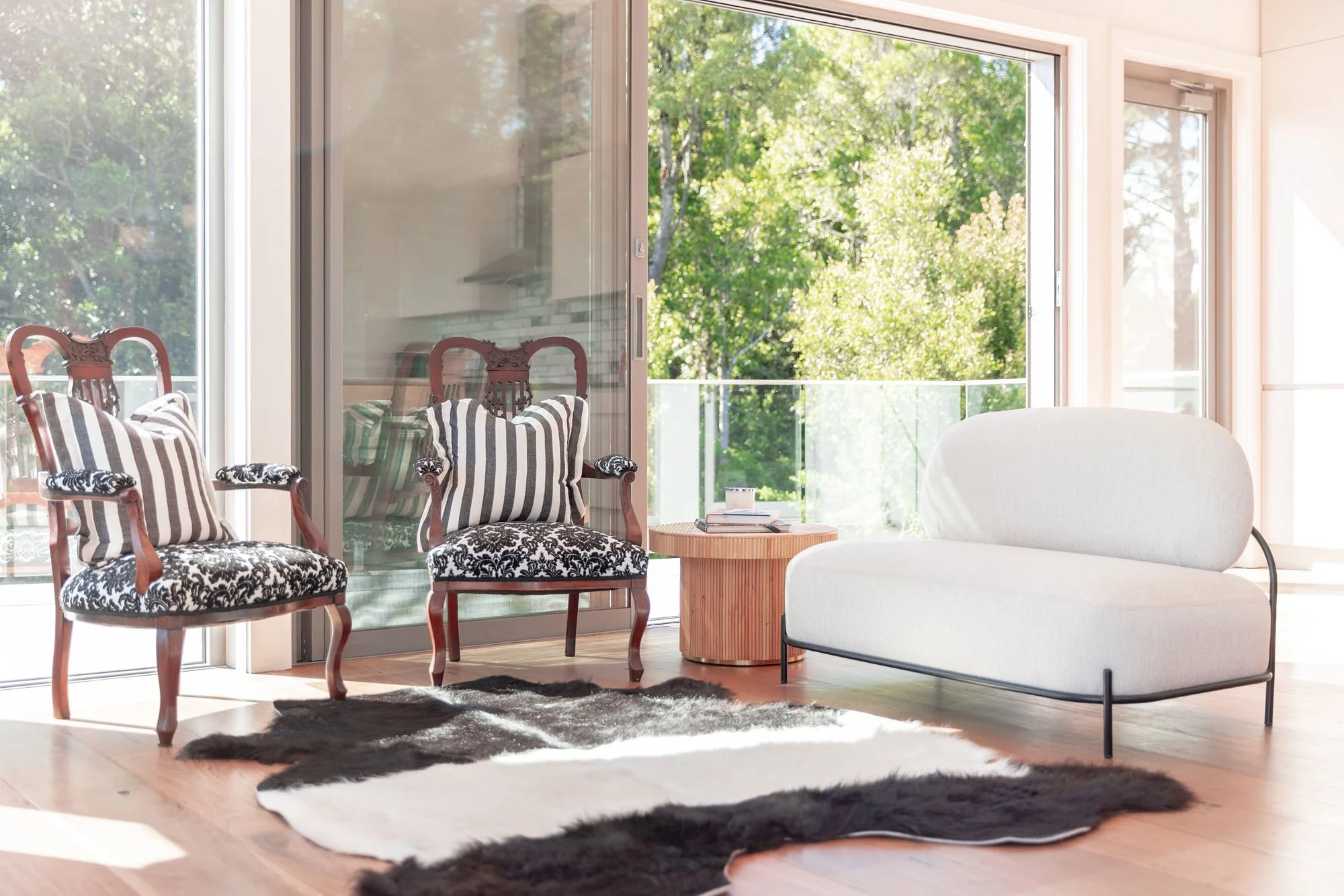 Living room with a white sofa, wooden side table, and vintage armchairs with black and white patterns, large windows with a view of green trees, black and white rugs, and sunlight filling the space.