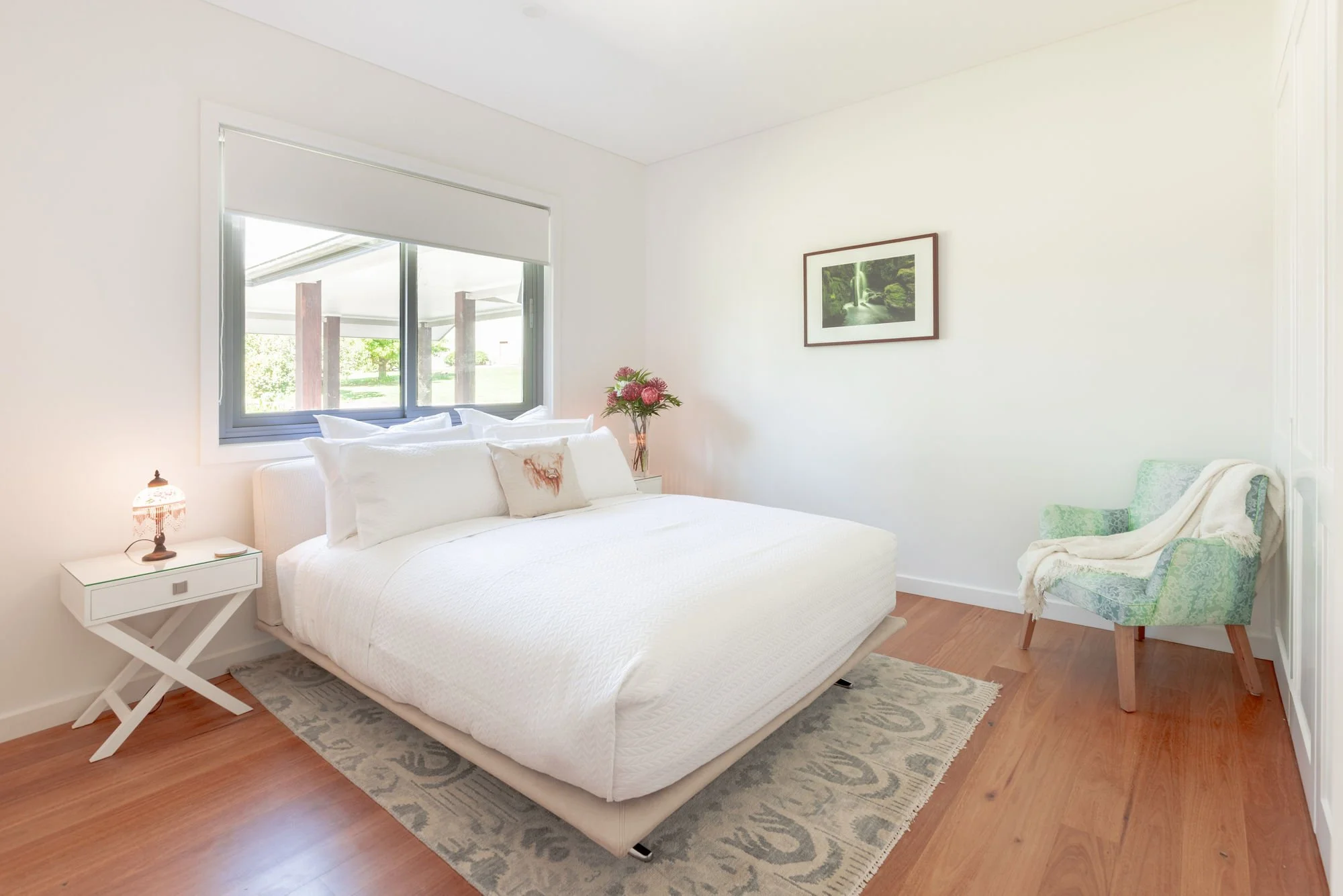 Bright bedroom with white bed, floral pillows, and a bedside table with a small lamp. A window shows an outside view of greenery and a covered porch. Wall has a framed nature photo, and there's a patterned armchair with a throw blanket.