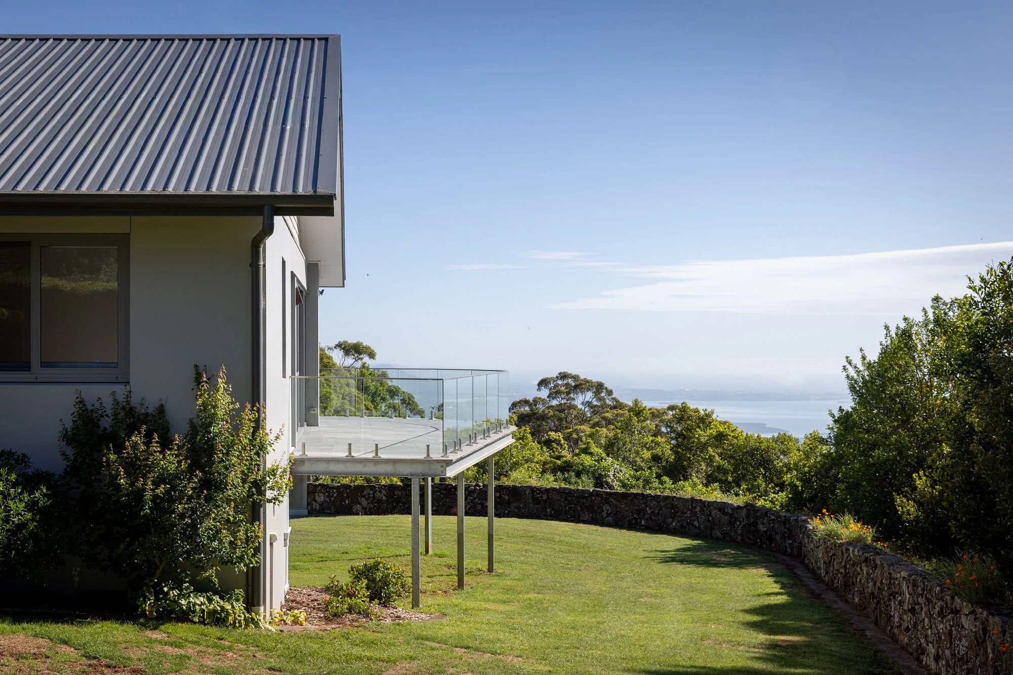 Modern white house with a metal roof and a glass balcony overlooking a lush green landscape with trees and water in the distance.