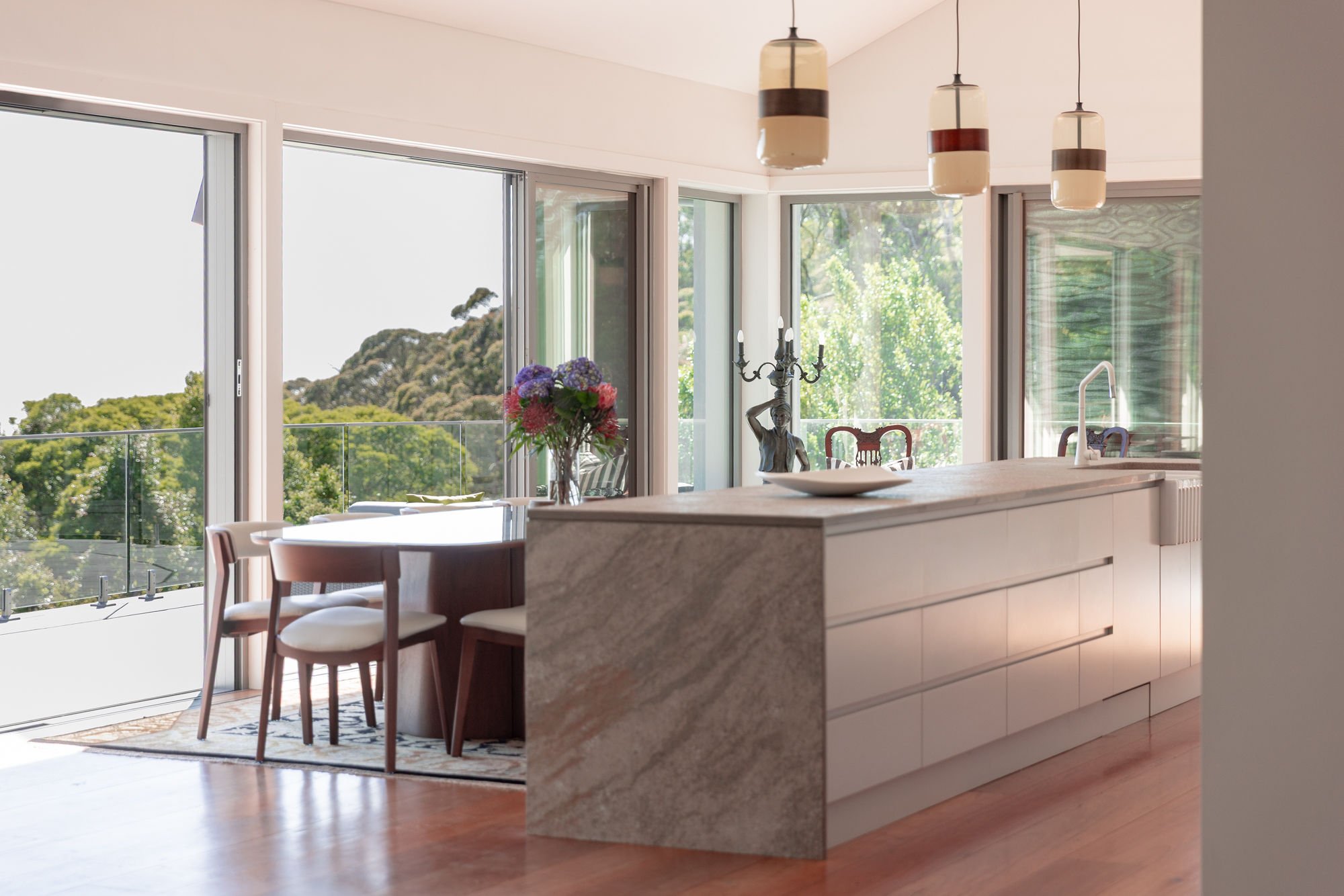Modern kitchen and dining area with large glass windows and doors, a marble island with a bowl and a vase of flowers, wooden chairs with white cushions, a chandelier, and a view of greenery and trees outside.