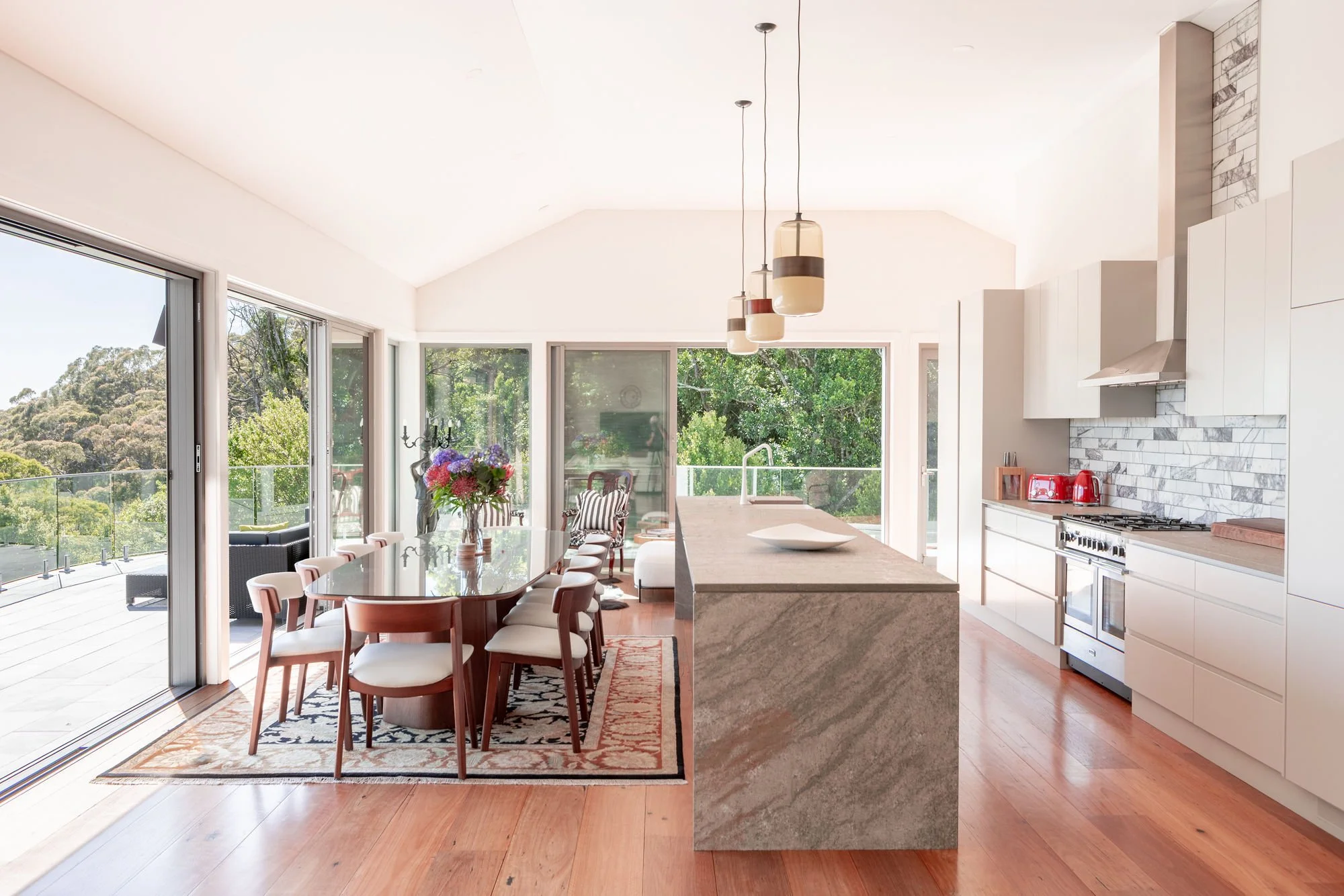 Bright kitchen and dining area with sliding glass doors opening to an outdoor deck with green trees. Interior features white cabinets, brick backsplash, modern light fixtures, and a wooden dining table with chairs, a flowers centerpiece, and a patter