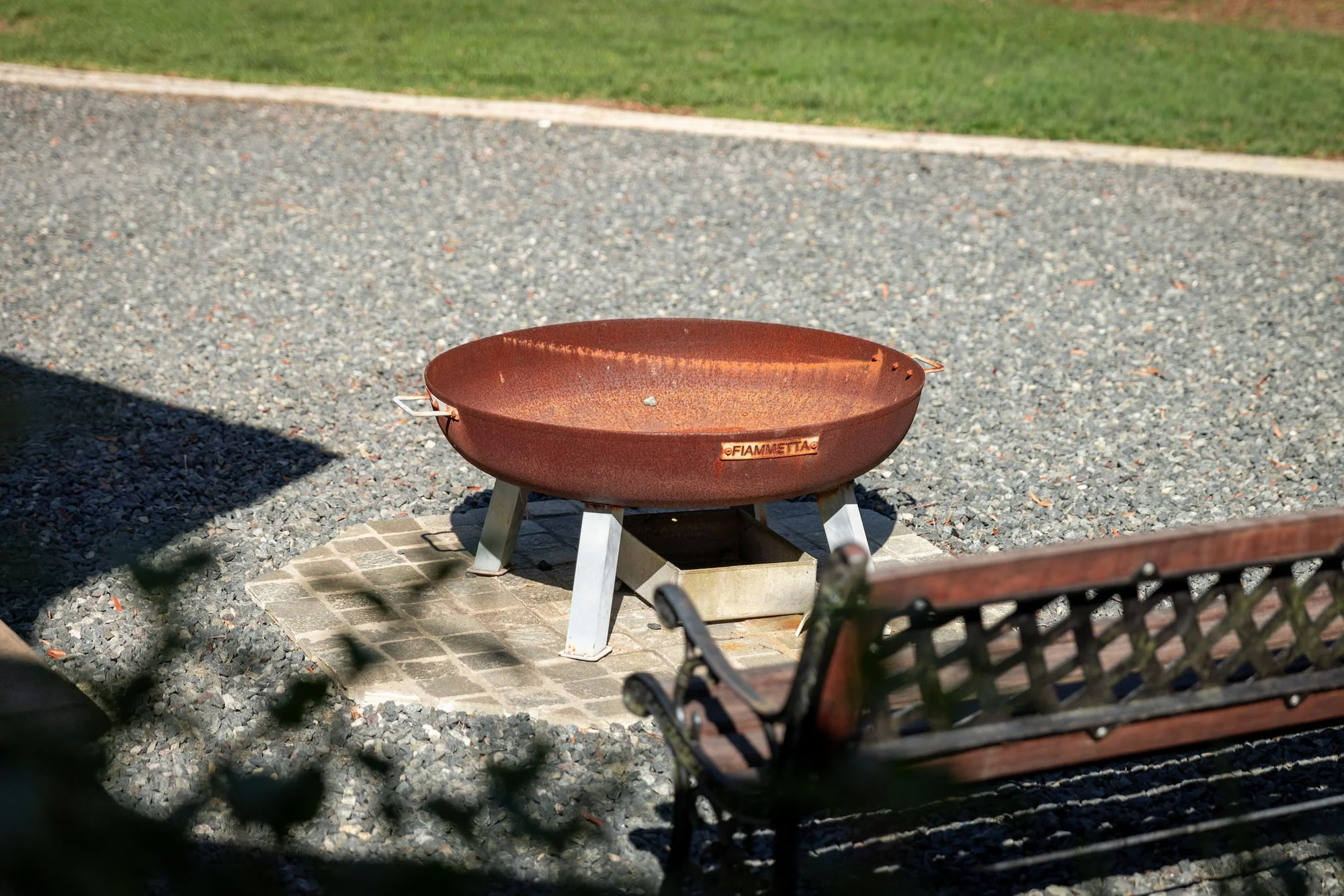 A rust-colored fire pit on a small brick platform, surrounded by gravel on a sunny day in a park or backyard with grass in the background.