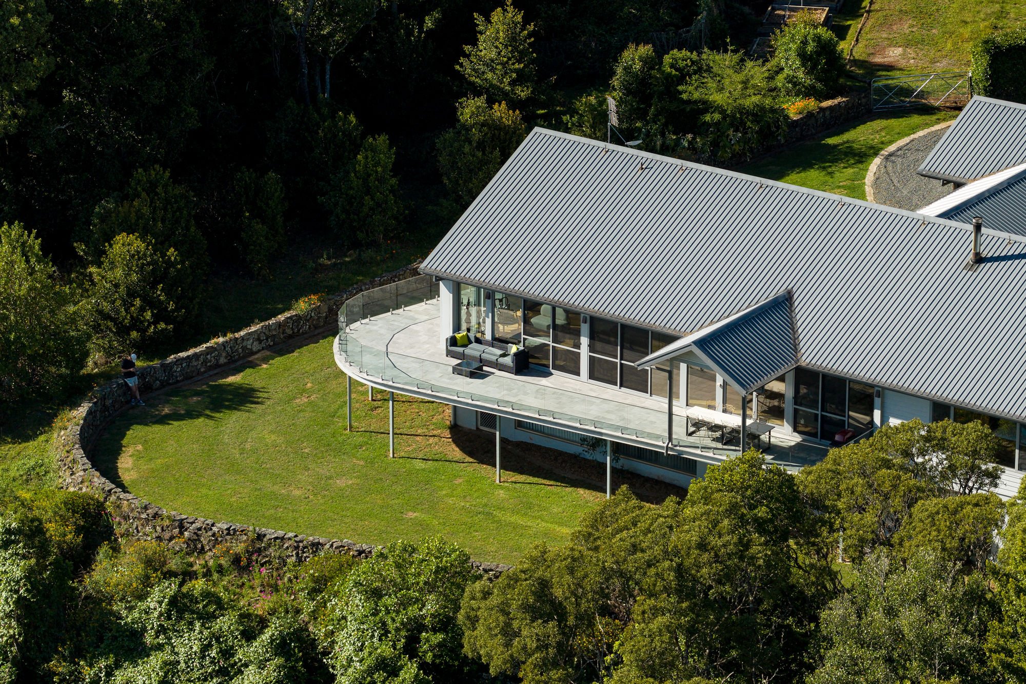 Aerial view of a modern house with a metal roof, large windows, and a curved balcony with outdoor furniture, surrounded by lush greenery and a stone wall.