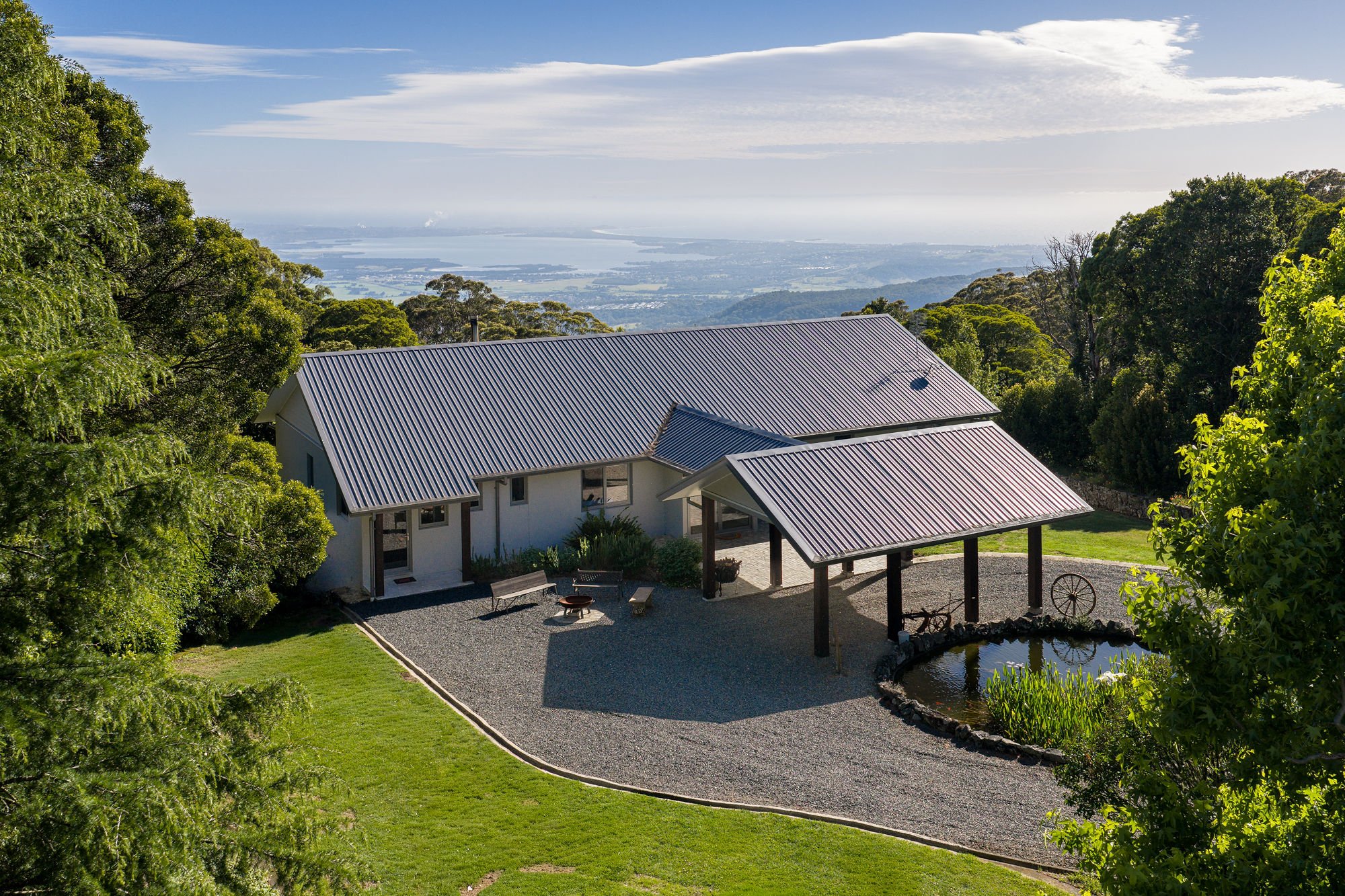 A house with a metal roof is surrounded by greenery and trees, overlooking a scenic view of an ocean or lake in the distance under a partly cloudy sky.