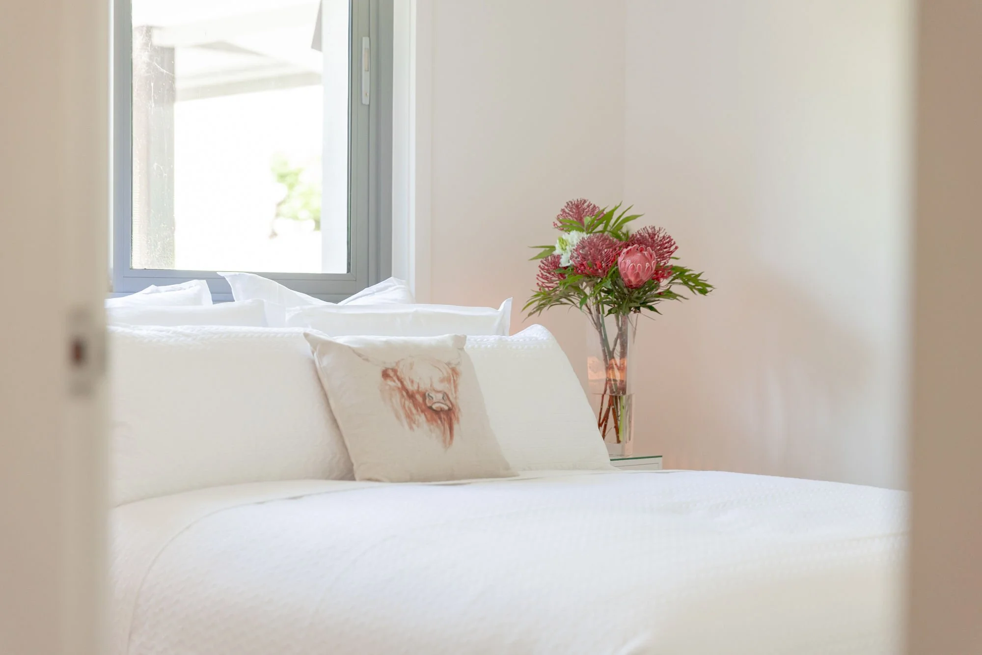 A bedroom with a white bed, white pillows, a throw pillow with an orange lion face, a window, and a vase of pink and red flowers on a side table or dresser.