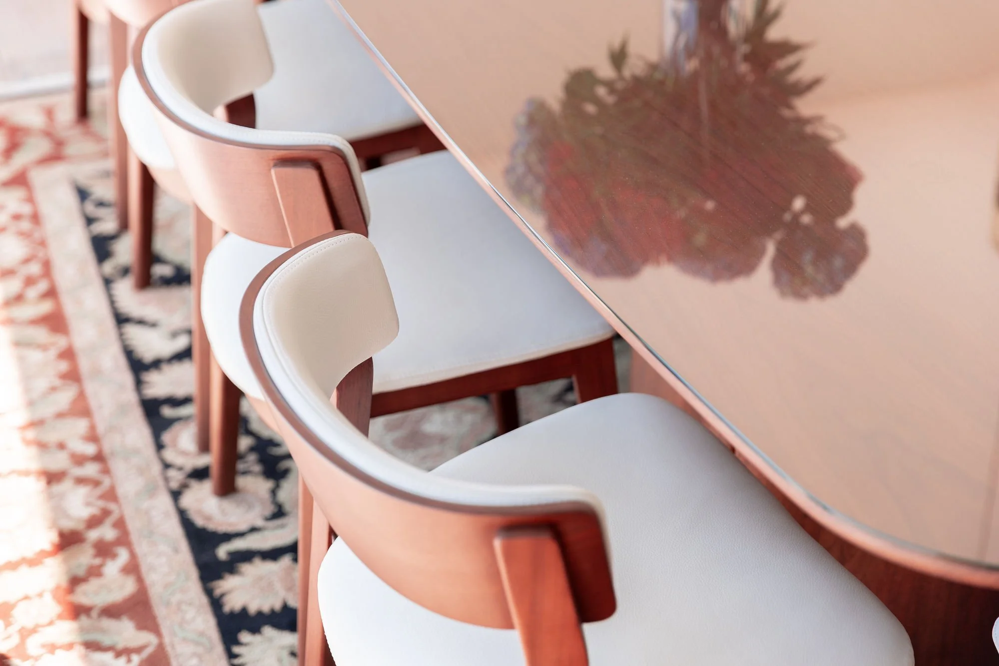 Close-up of a peach-colored dining table with a reflective surface showing a tree's reflection, surrounded by matching chairs with cream-colored cushions, on a patterned rug.