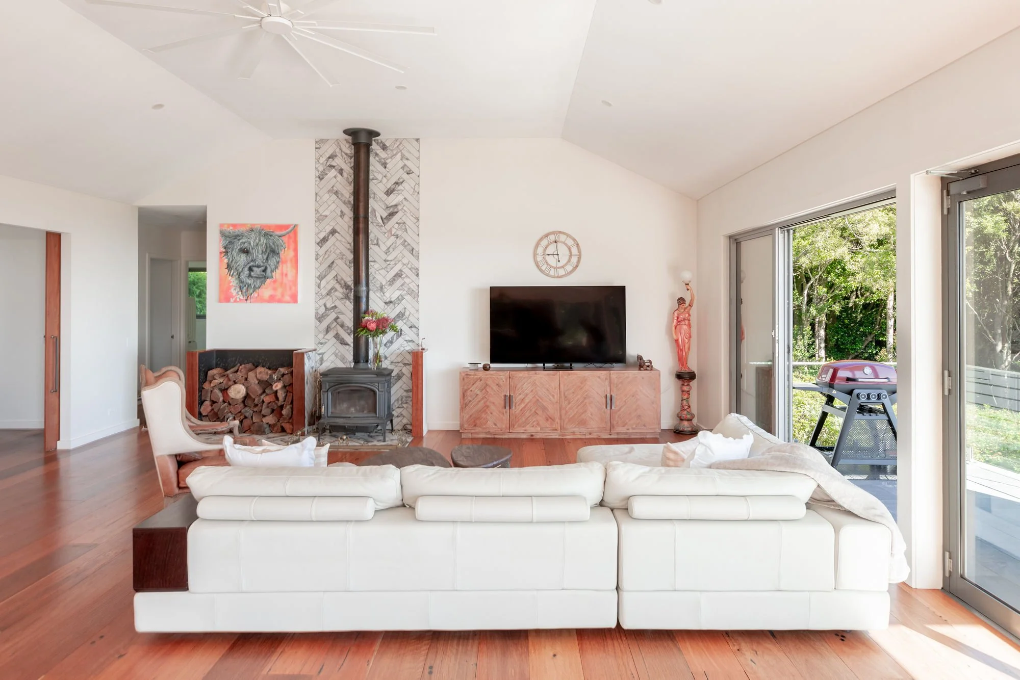 Modern living room with white sofa, wood stove, fireplace logs, artwork, and large sliding glass doors leading outside to greenery.