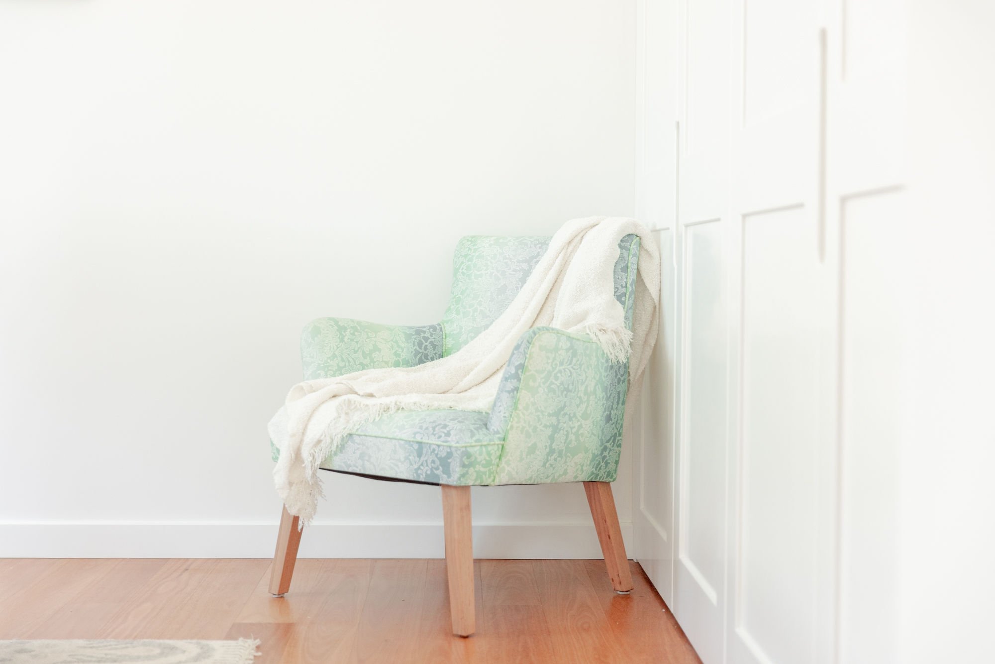 A green upholstered armchair with a white throw blanket draped over it, positioned against a white wall in a room with hardwood floors.