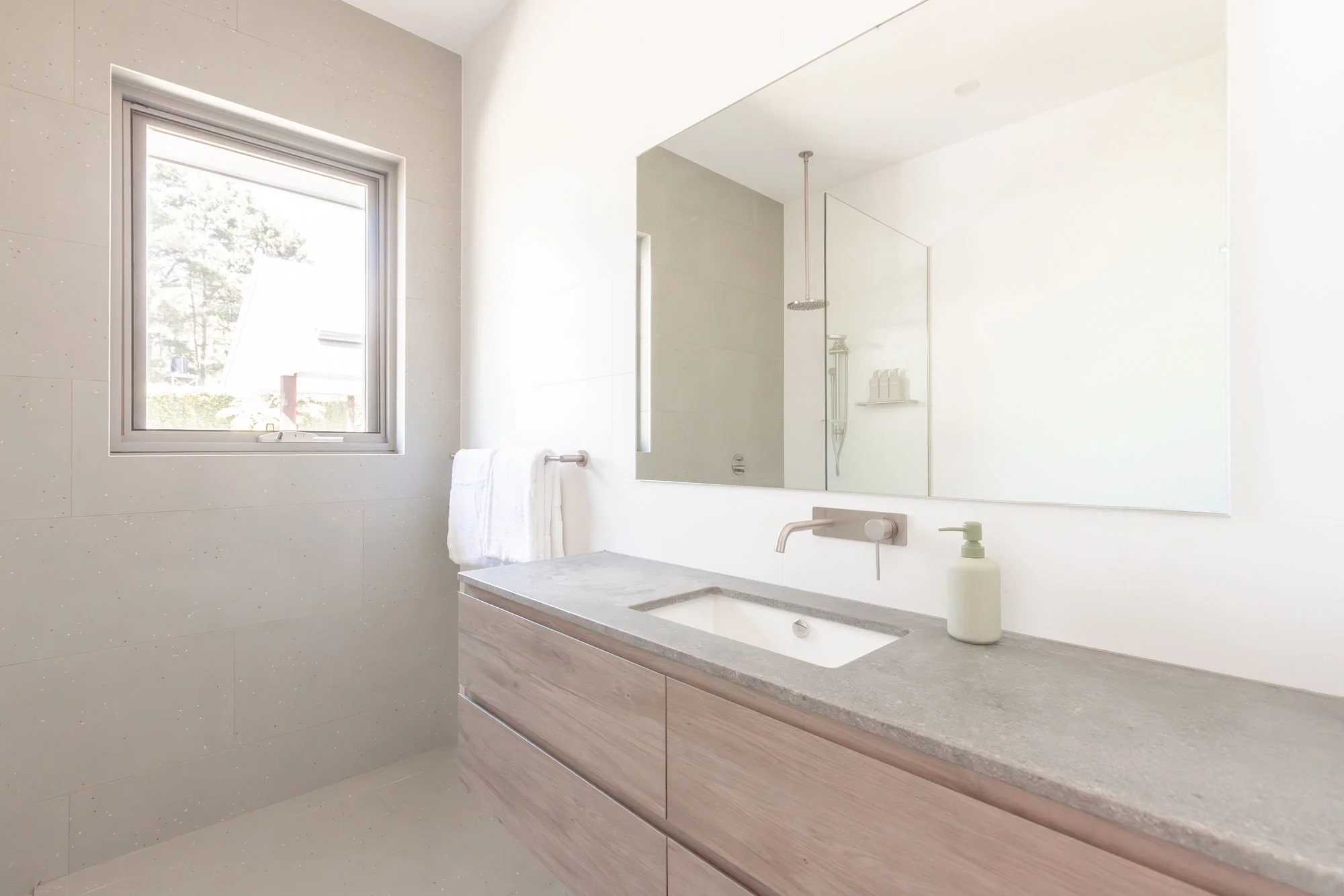 Minimalist bathroom with a large mirror, a sink with a wooden cabinet, a white soap dispenser, a window letting in natural light, and a towel hanging on a rail.