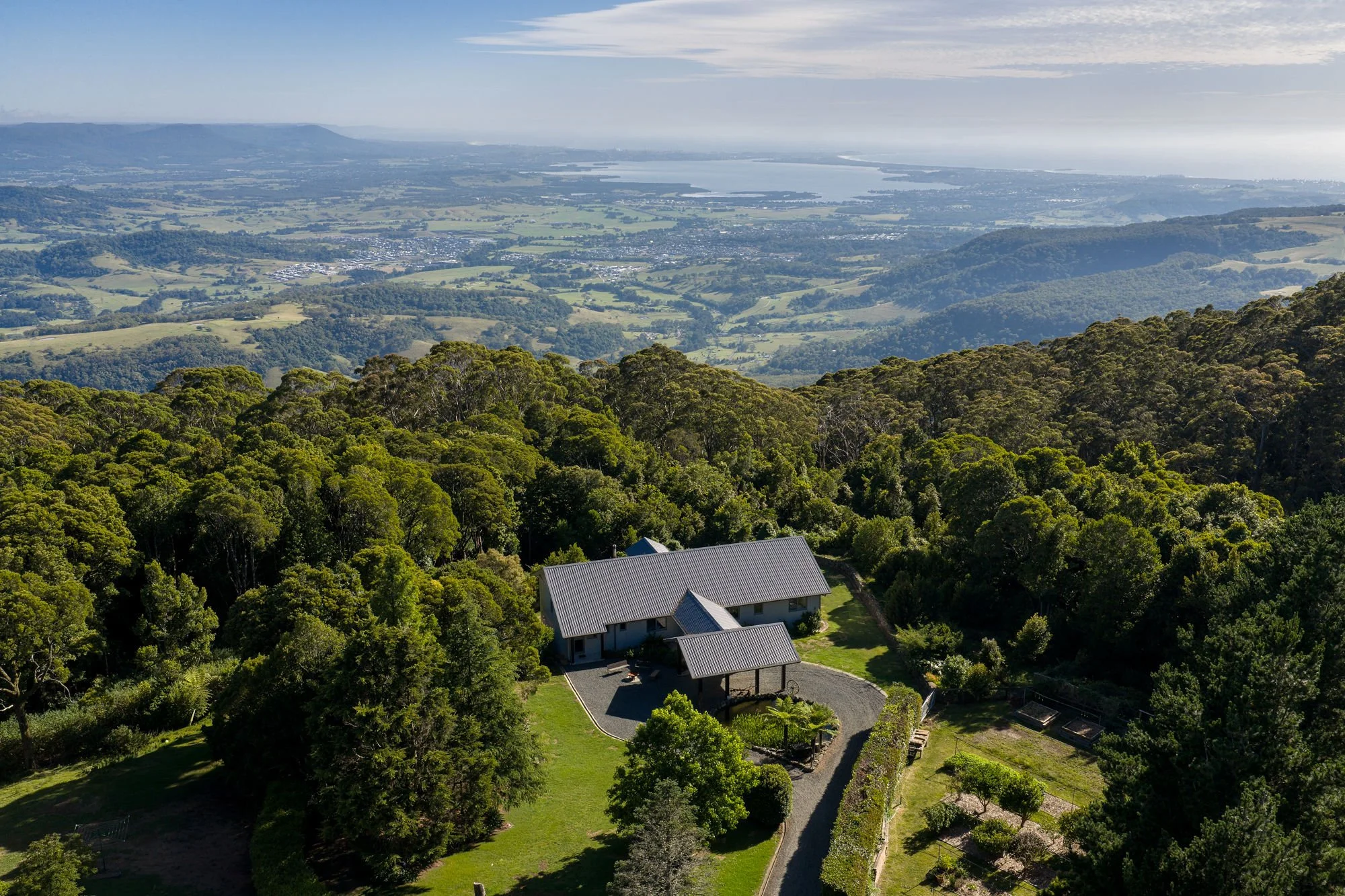 Aerial view of a house surrounded by lush green trees, with a winding driveway leading to the house, set in a hilly landscape with a distant body of water and mountains in the background.