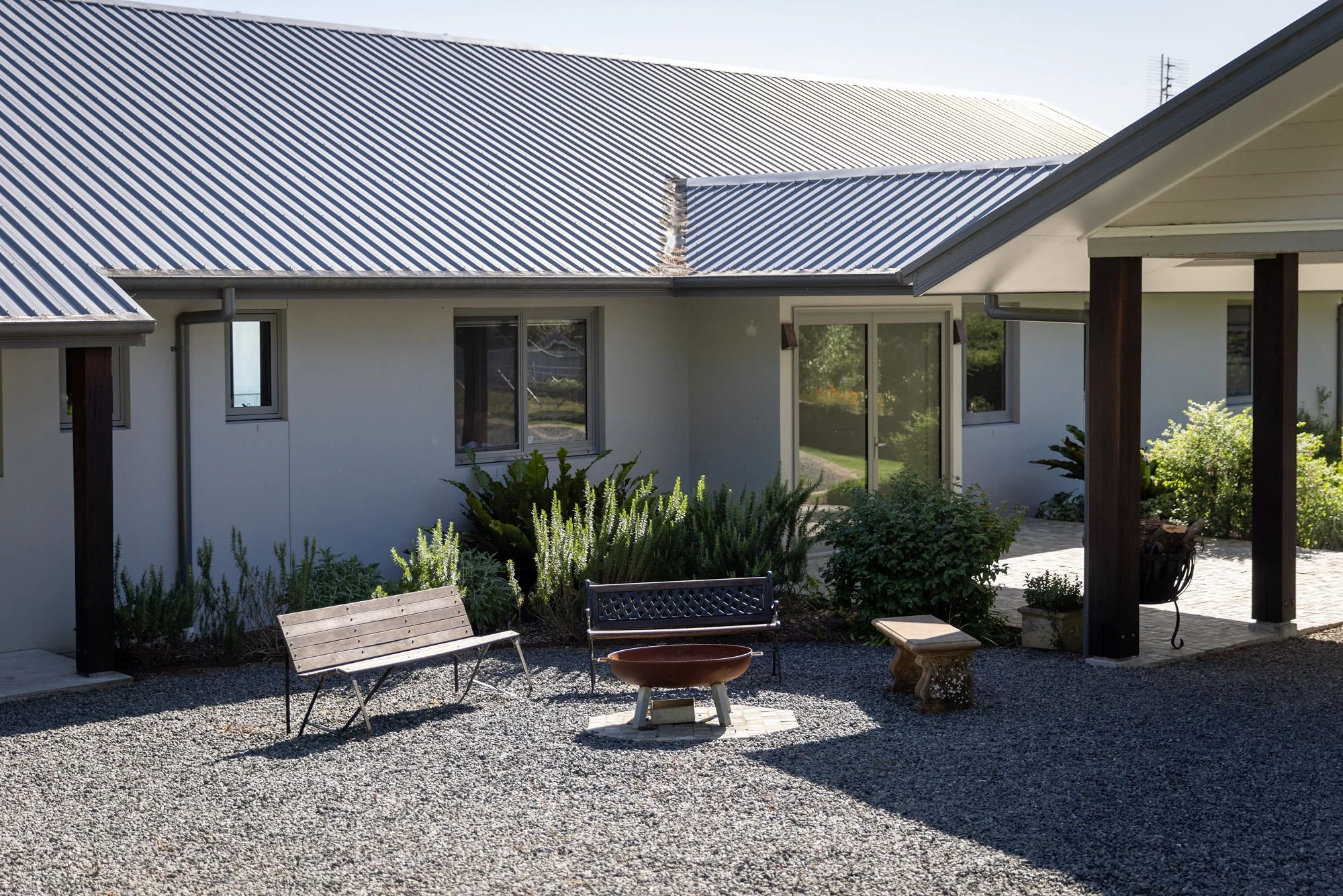 A residential backyard with a gravel ground, garden plants, a fire pit, and a modern house with a metal roof and large windows.