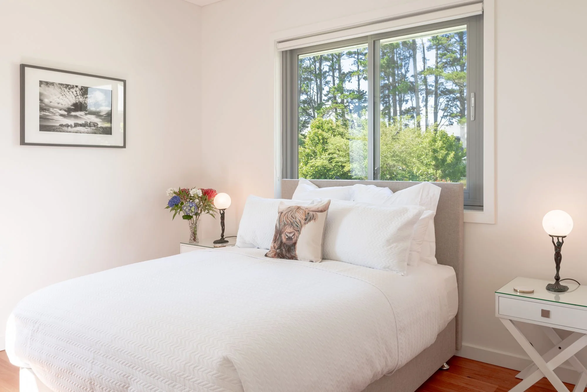 A bedroom with a large white bed featuring a decorative pillow with a lion's face, beside a white nightstand with a glass vase of colorful flowers and a lamp. There are two nightstands, one on each side of the bed, with matching lamps. A picture in b