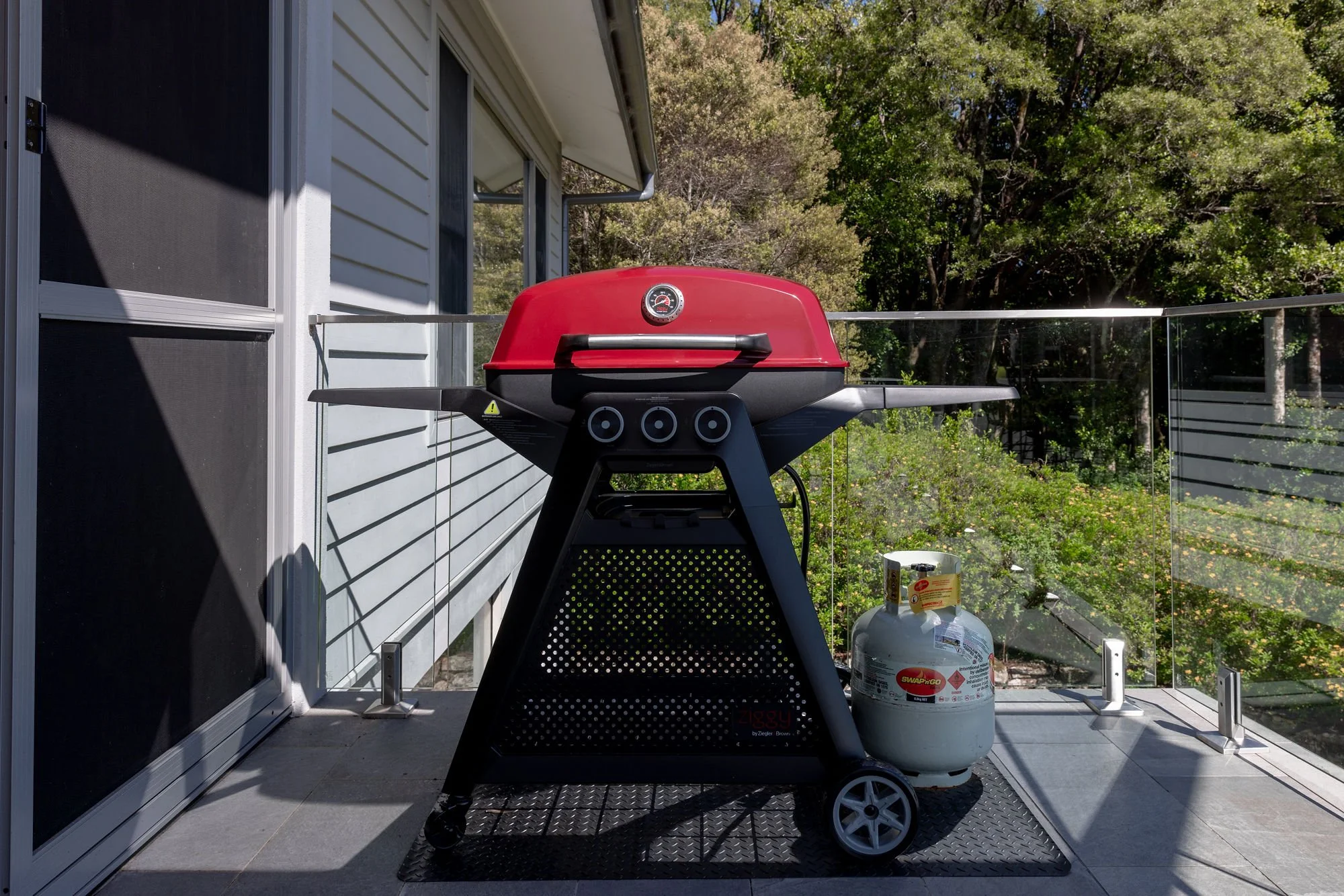 Red gas grill with black stand on a balcony, with propane tank underneath, surrounded by glass railing and trees in the background.