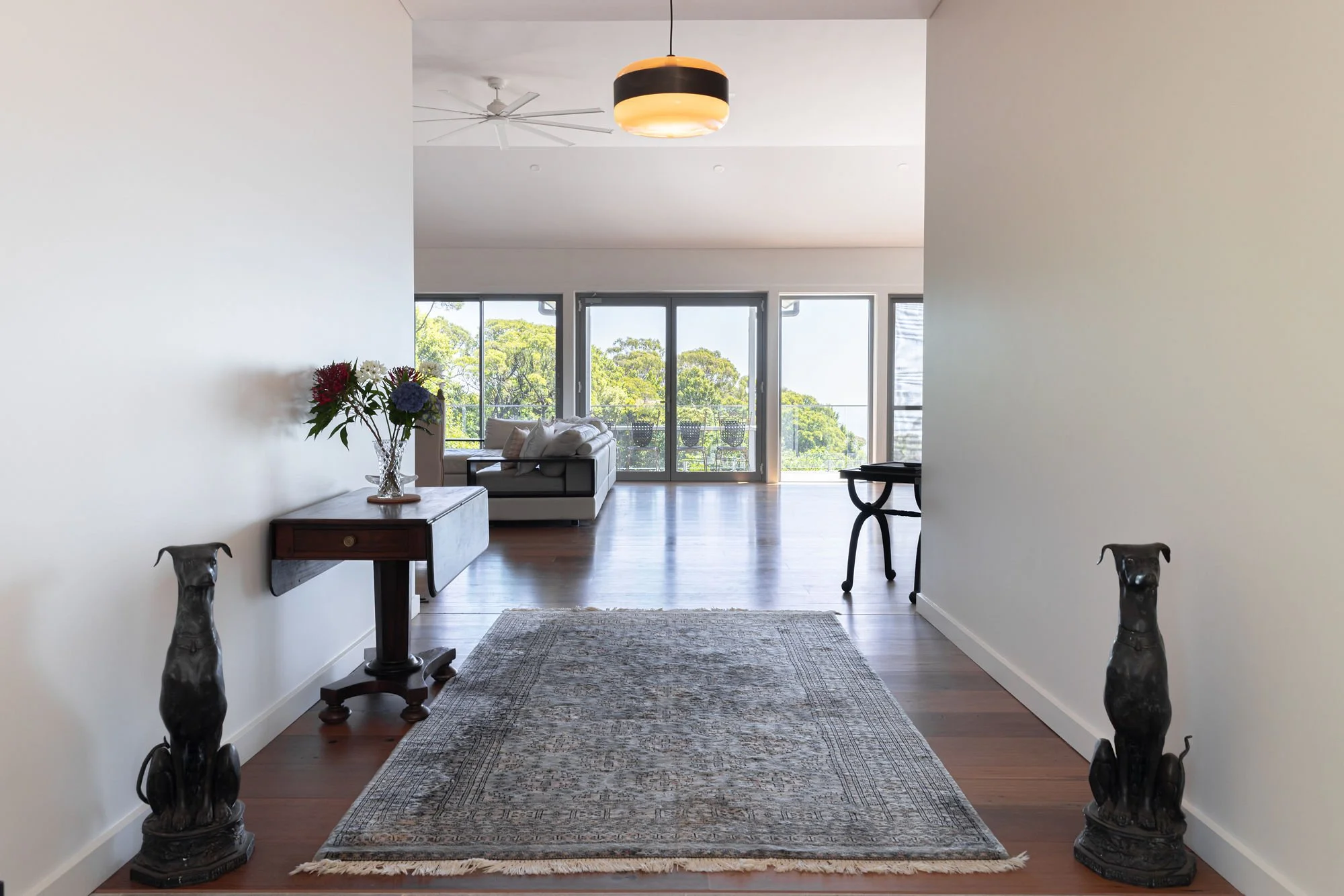 View of a modern living room seen from an entryway, with white walls, a patterned area rug, and decorative elephant statues flanking an entry table. The room has large glass sliding doors leading to a balcony with outdoor chairs, overlooking lush gre