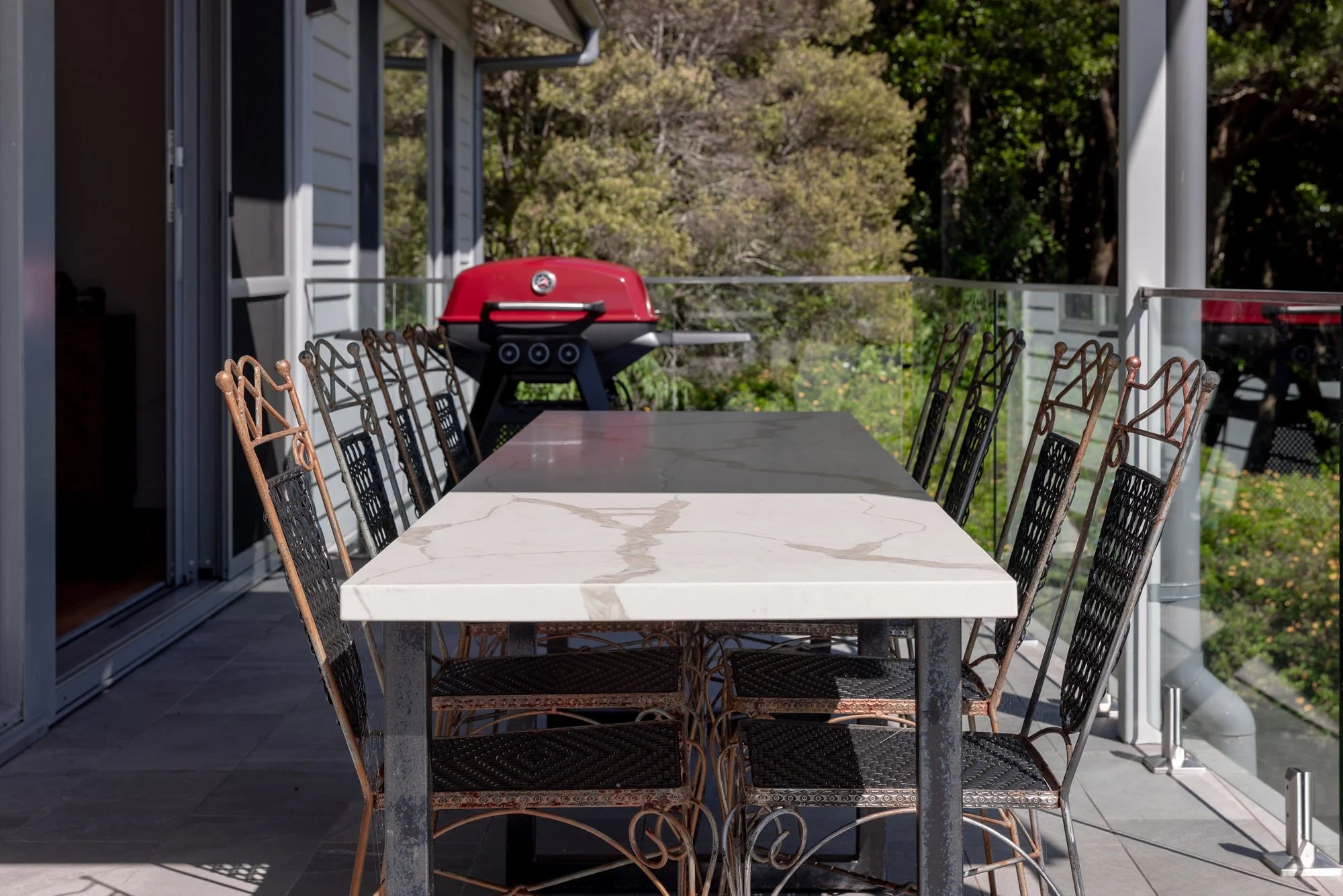 Outdoor balcony with a long white marble table, eight vintage-style chairs, and a red portable grill, overlooking lush green trees.