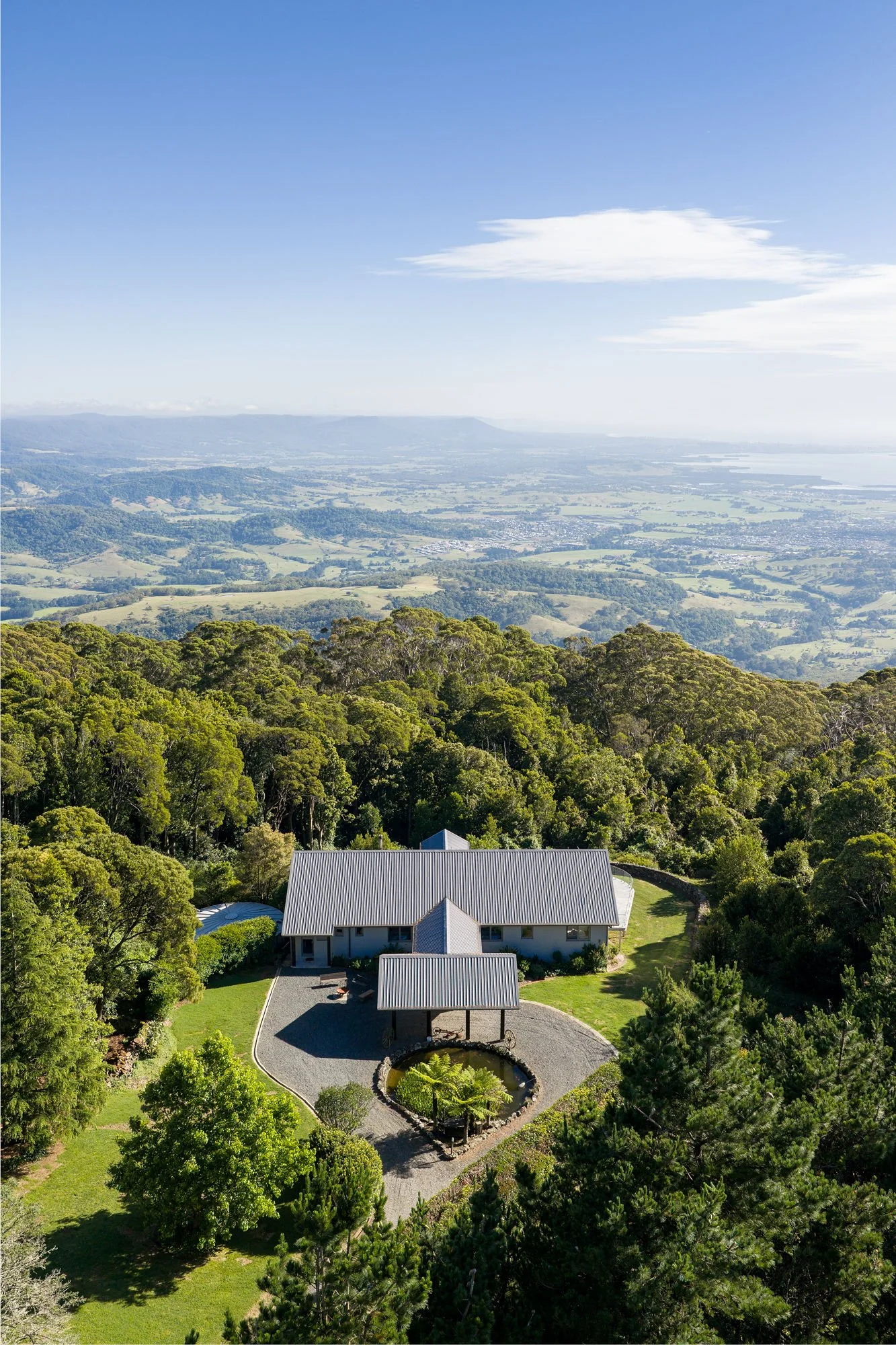 A scenic view of a house surrounded by lush green trees and a pond, with rolling hills and a distant landscape under a clear blue sky with some clouds.