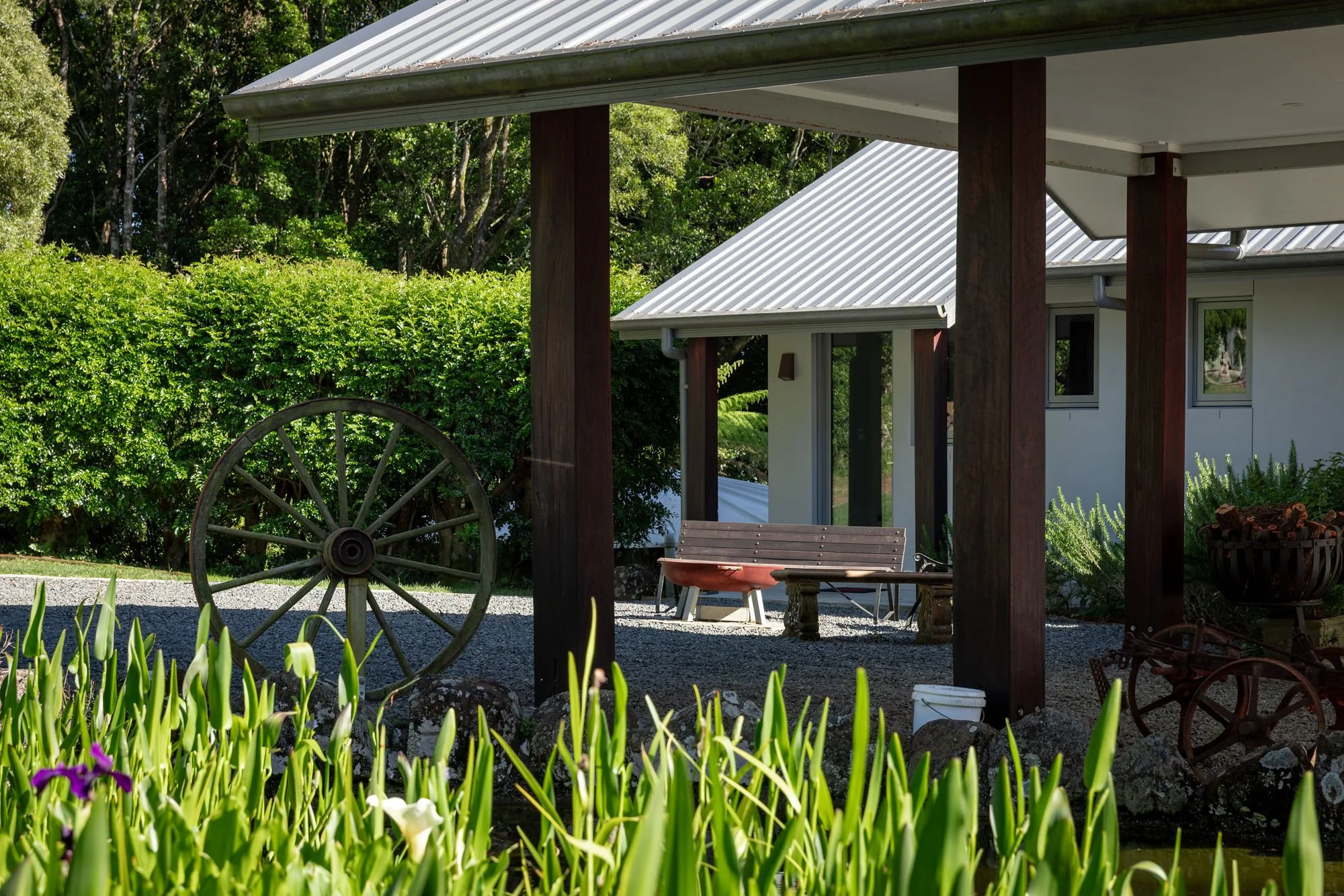 A backyard patio with a wooden bench, a wagon wheel, potted plants, and a white house with a metal roof surrounded by greenery and flowering plants.