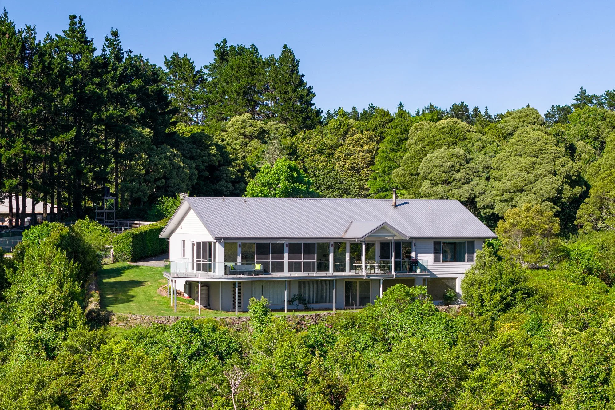 A white house with a gray metal roof surrounded by lush green trees and a well-maintained lawn under a clear blue sky.
