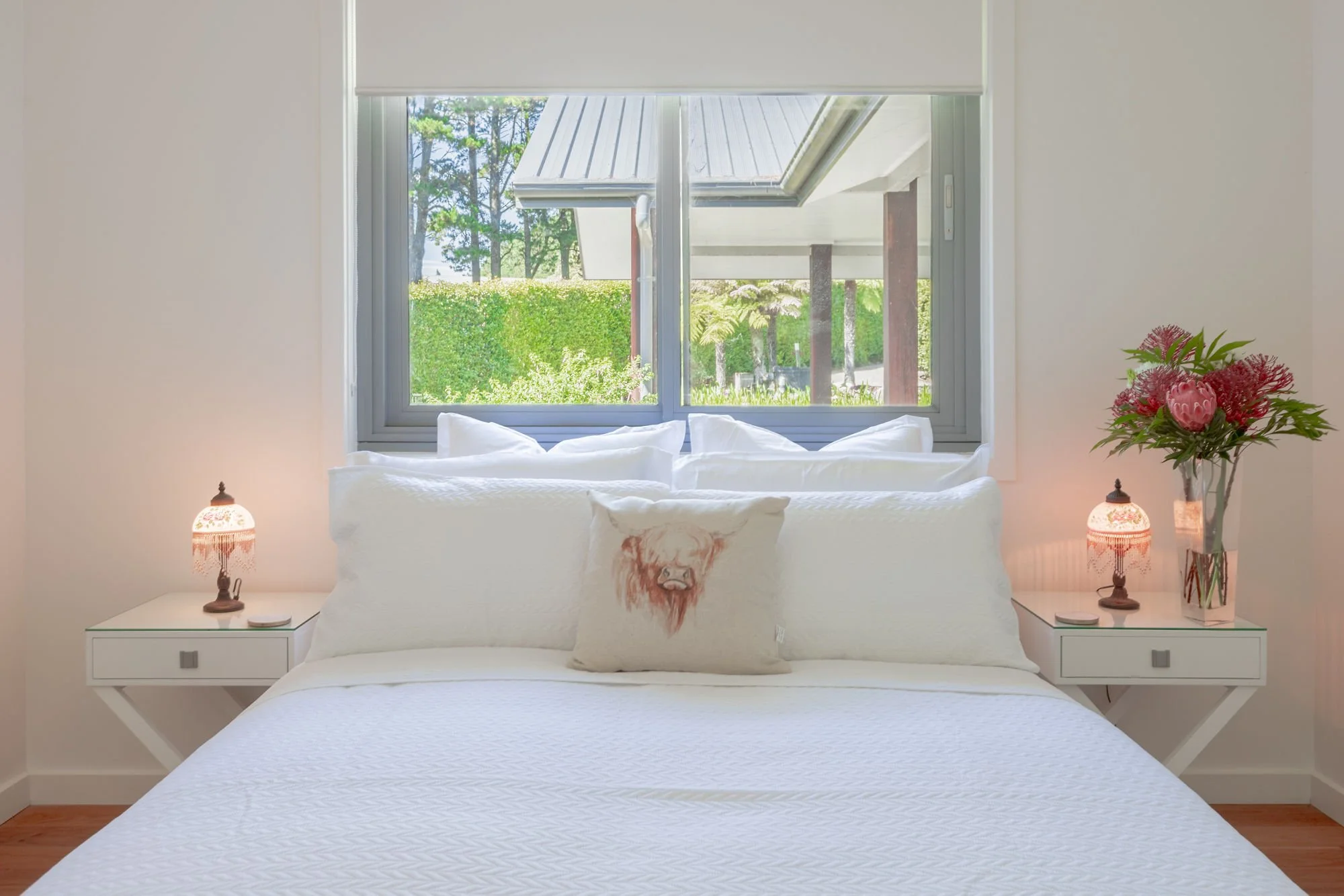 A bedroom with a large window showing trees outside, white walls, and a white bed with a pillow featuring a lion's face. On each side of the bed are nightstands with decorative lamps, and a vase with pink and red flowers is on the right nightstand.