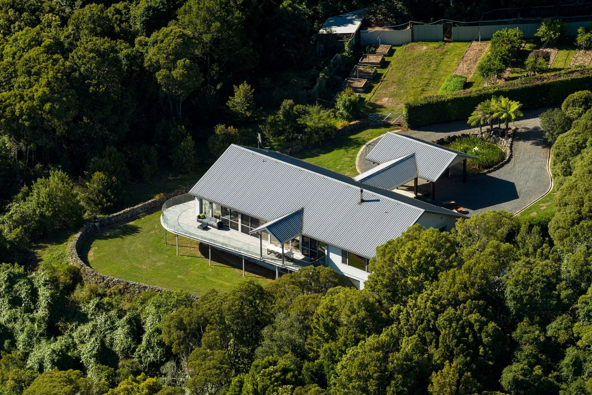 Aerial view of a house with a metal roof surrounded by lush trees, a garden area, and a gravel driveway.