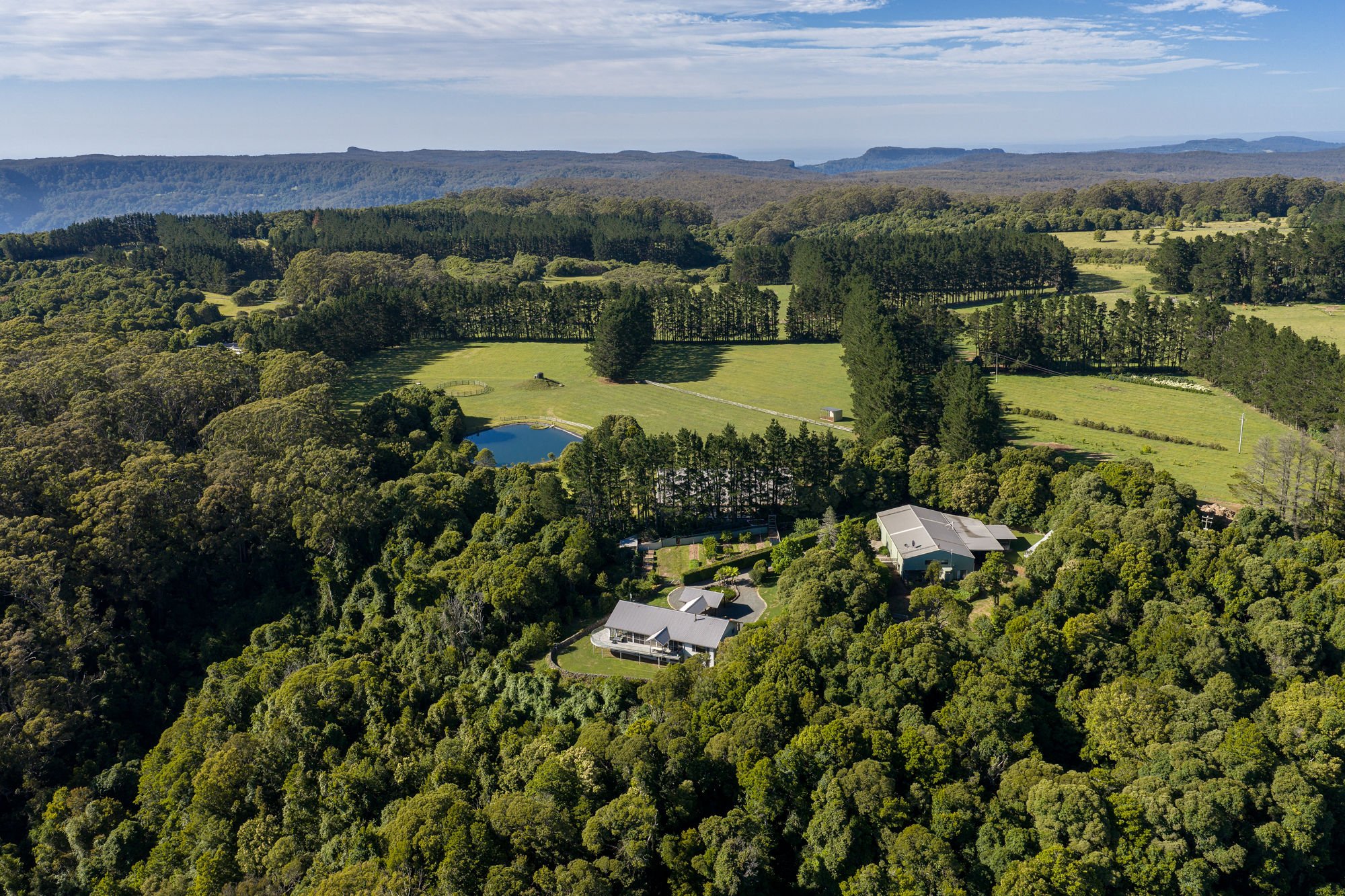 Aerial view of a lush green landscape with trees, a small pond, fields, and a few buildings under a partly cloudy blue sky.