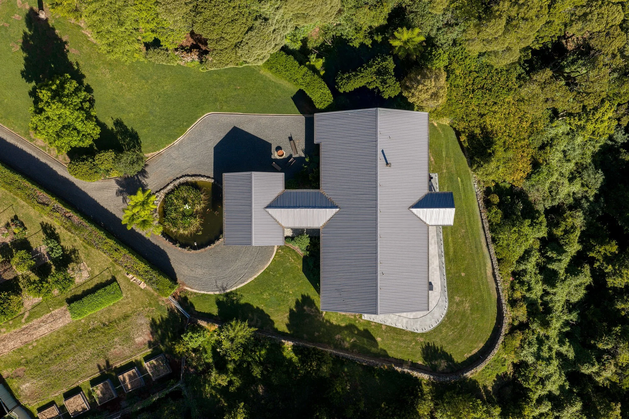 An aerial view of a house with a grey metal roof, surrounded by trees and green lawns, with a gravel driveway leading to the house.