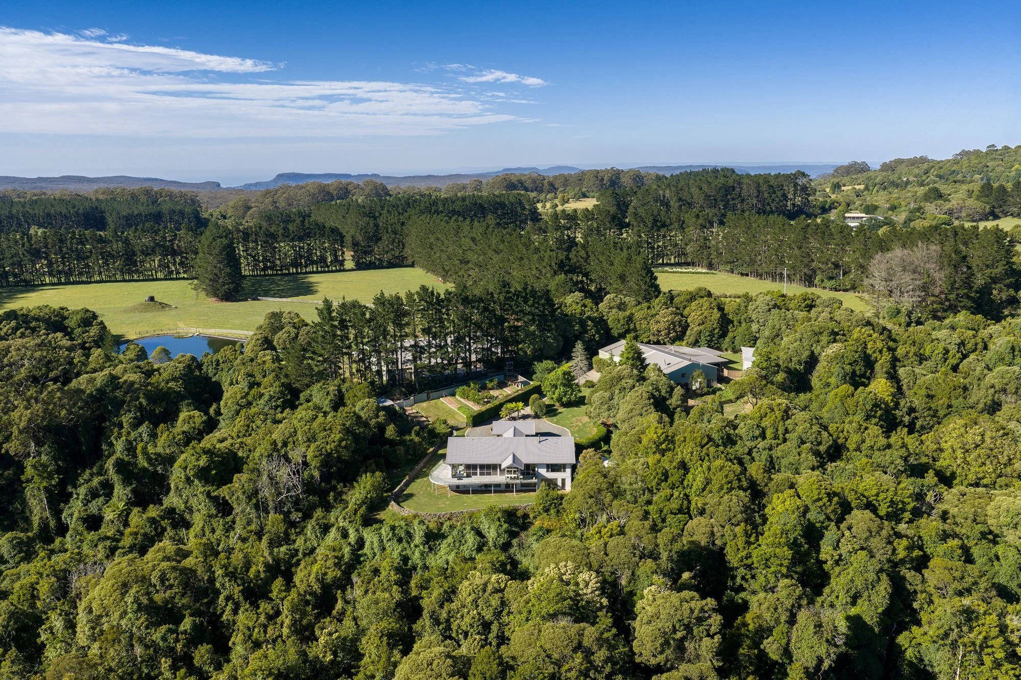Aerial view of a house in a lush green forest with rolling hills in the distance and a partly cloudy blue sky.