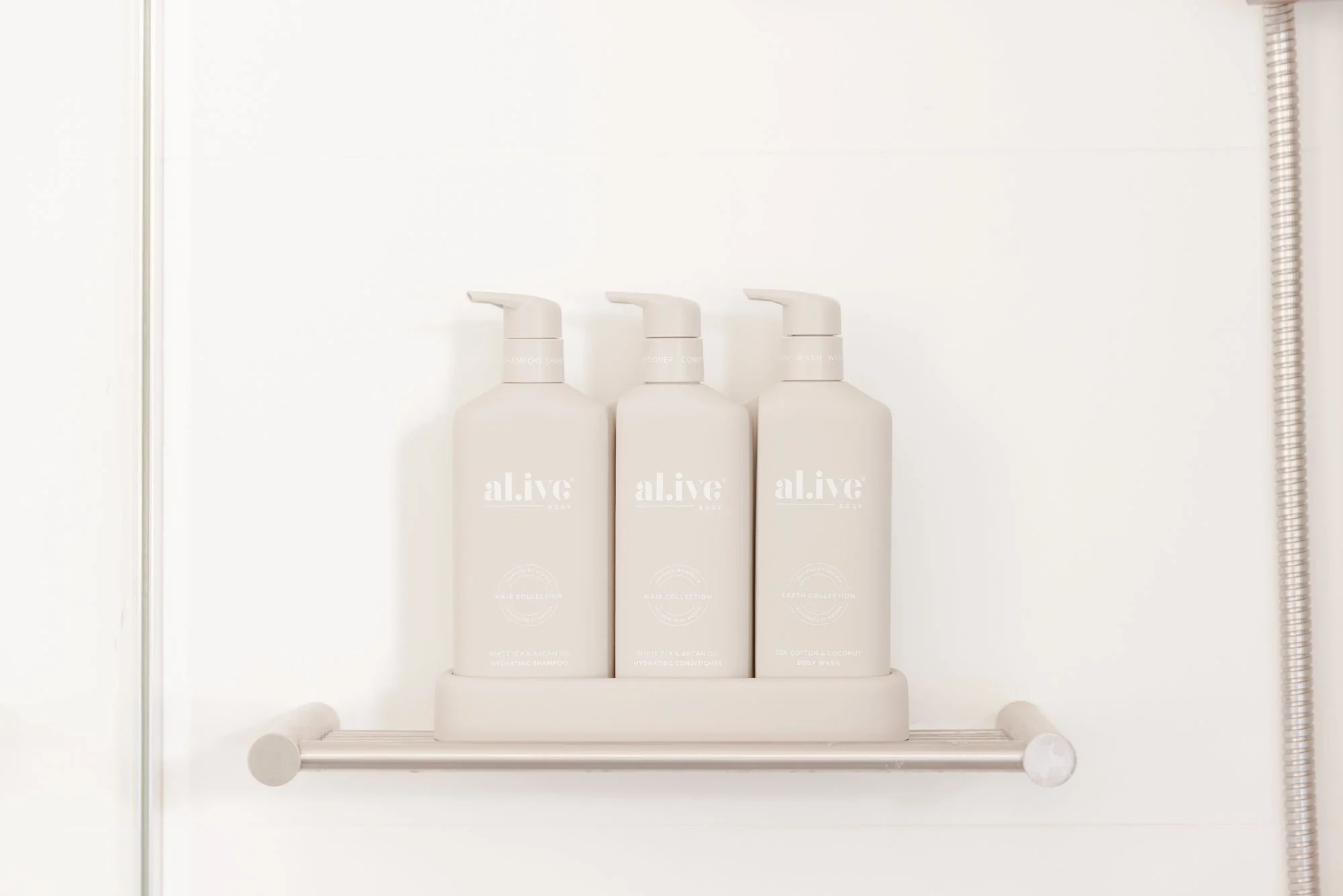 Three white bottles of personal care products on a bathroom shelf with a white background. The bottles are labeled 'a.live' and include shampoo, conditioner, and body wash.