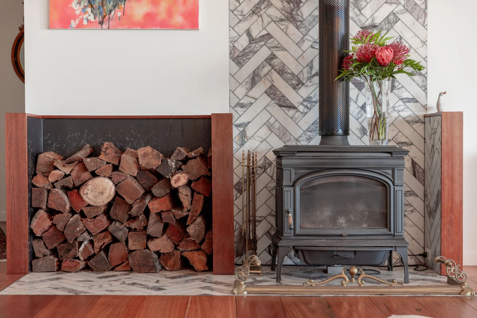 Firewood stored in a fireplace with a black mantel, next to a grey stove, and a vase with pink flowers on a marble wall.
