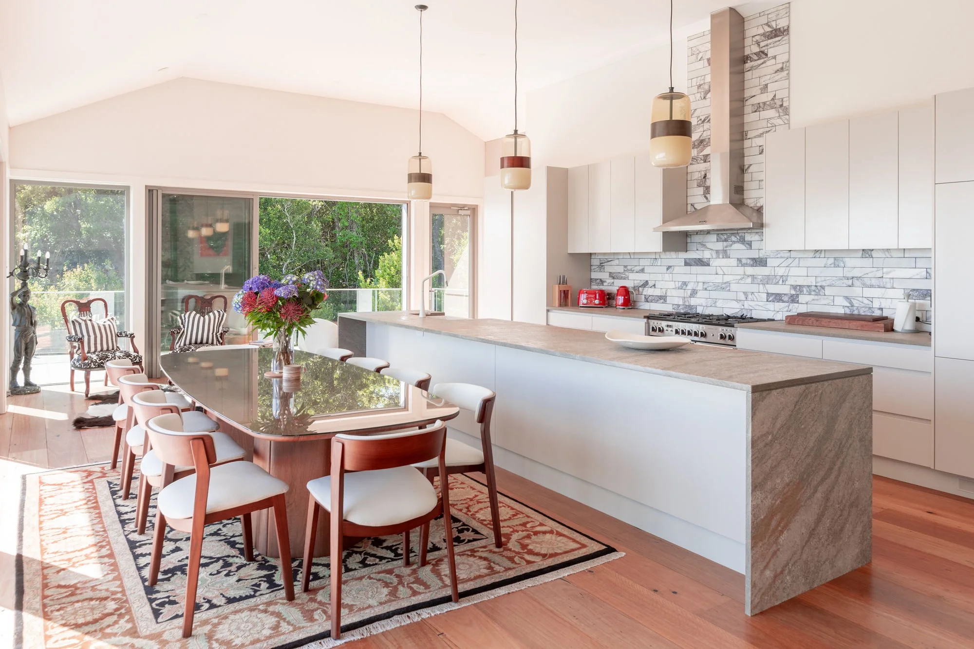 Modern open-concept kitchen and dining area with large window, wooden flooring, white cabinetry, marble backsplash, and a dining table with wooden chairs. Decor includes a vase of flowers and pendant lights.