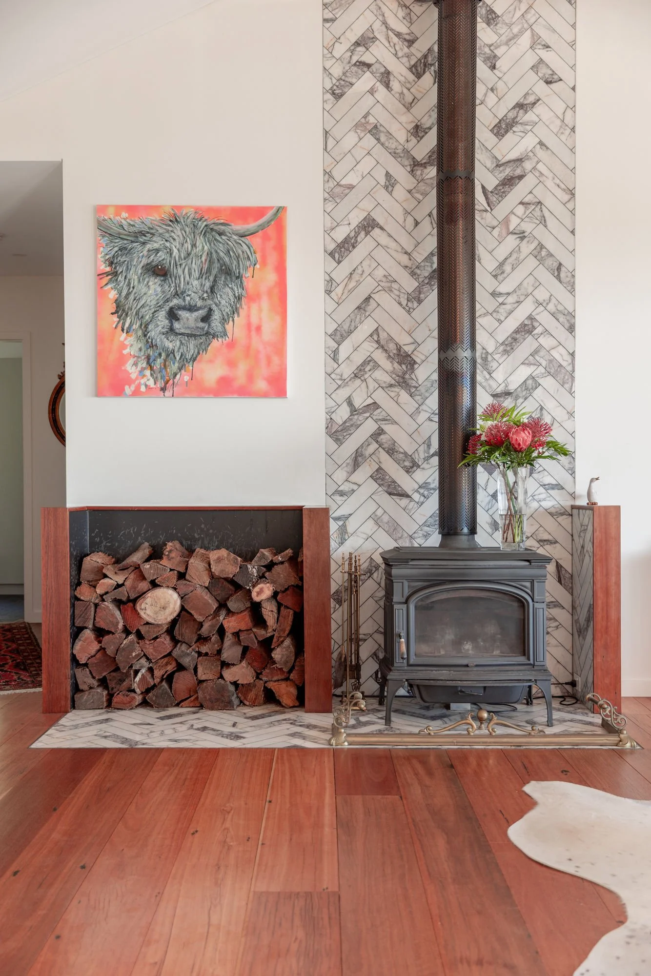 Living room corner with a stacked firewood storage, a painting of a Highland cow on the wall, a wood stove, a vase of pink flowers, and a decorative white cow sculpture, all set against a herringbone marble wall and hardwood floor.