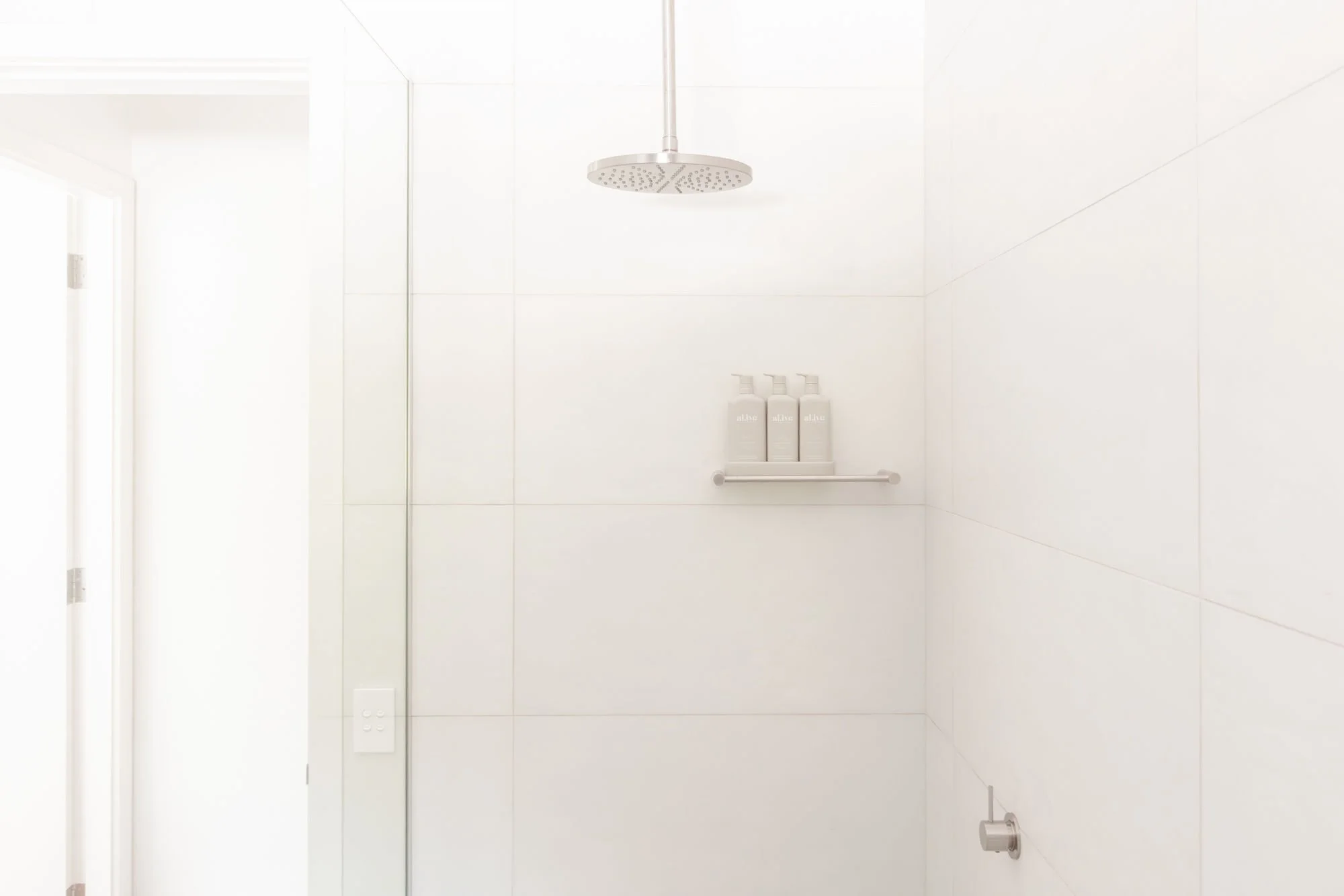 Shower with a rain showerhead, three bottles on a shelf, white tiled walls and a glass door.