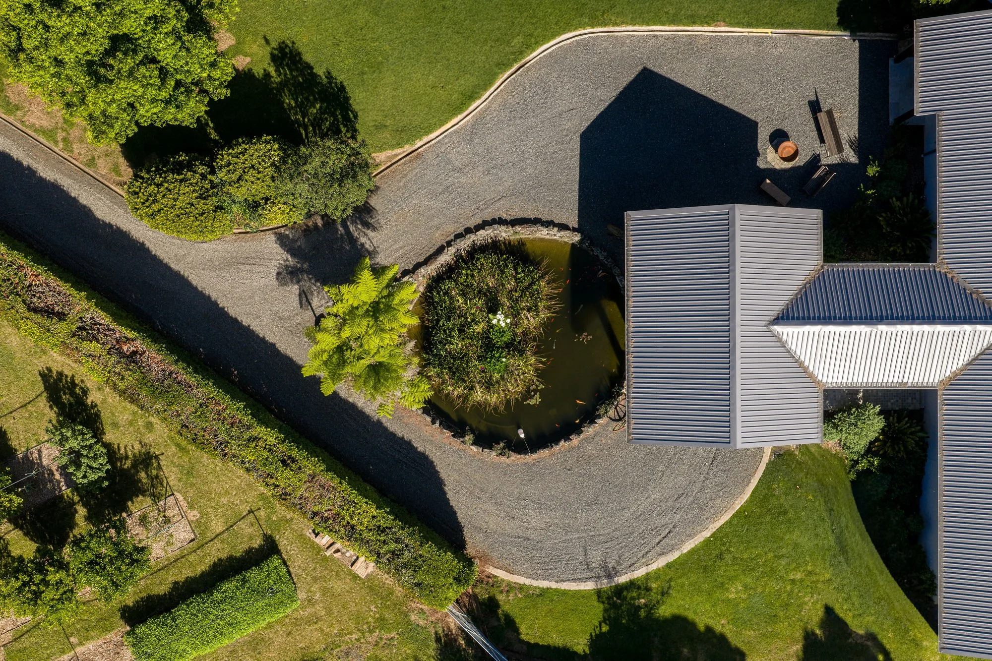 An aerial view of a residential backyard with a small pond, trees, a gravel pathway, two buildings with metal roofs, and outdoor furniture.