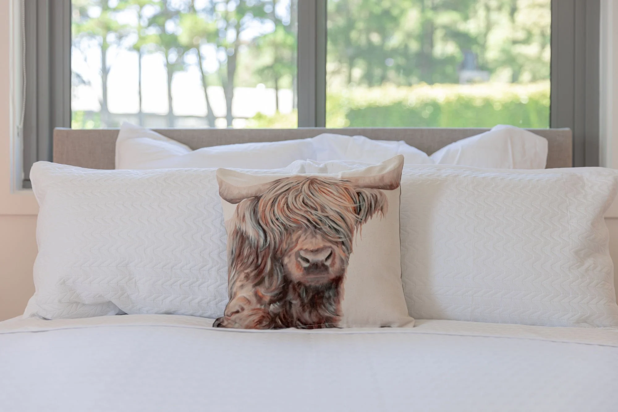 Decorative pillow with a high-resolution image of a dog’s face placed on a bed with white bedding, in front of a large window showing green trees outside.