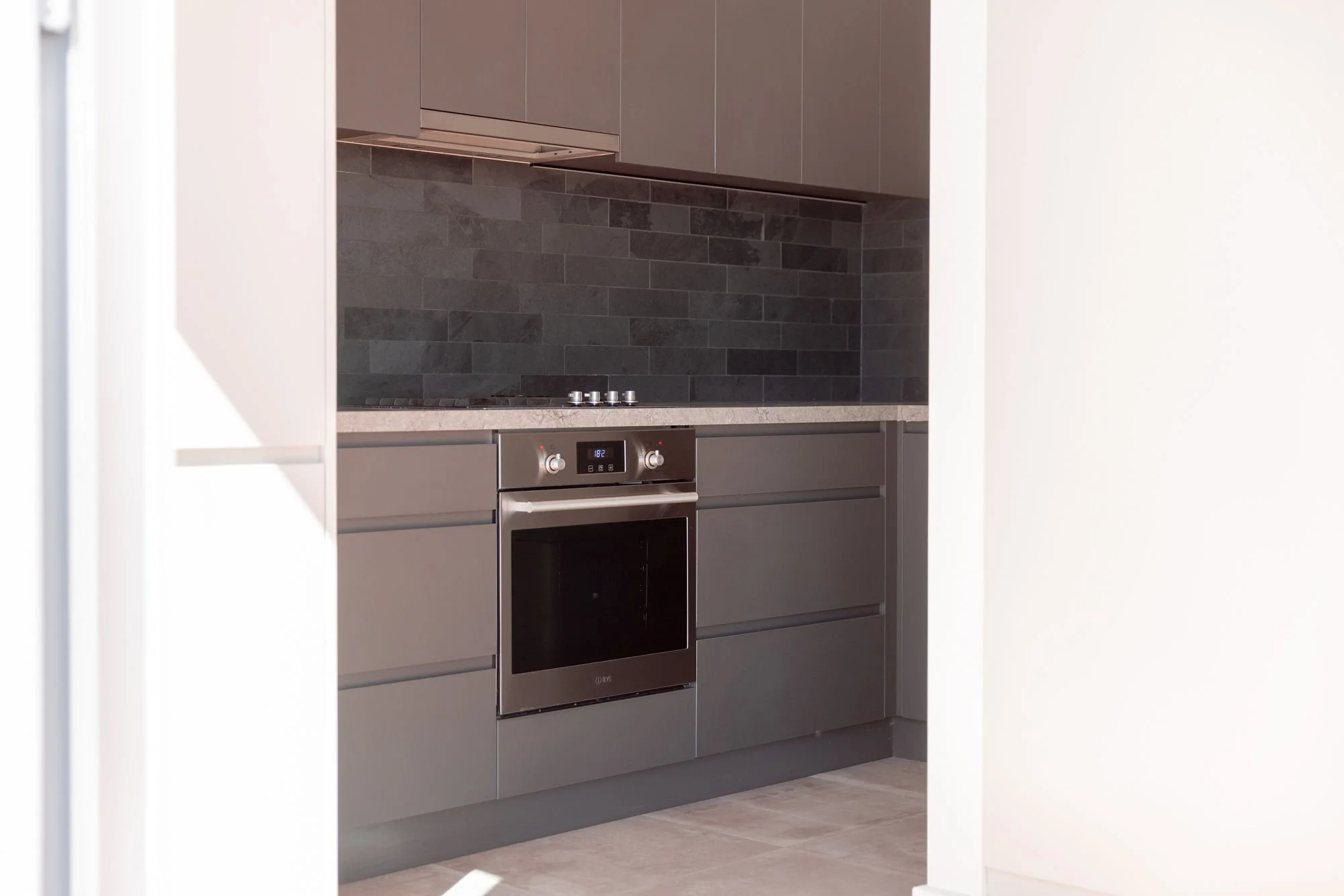 Modern kitchen with grey cabinets, black tiled backsplash, stainless steel oven, and a cooktop. Part of the room is visible through a doorway.