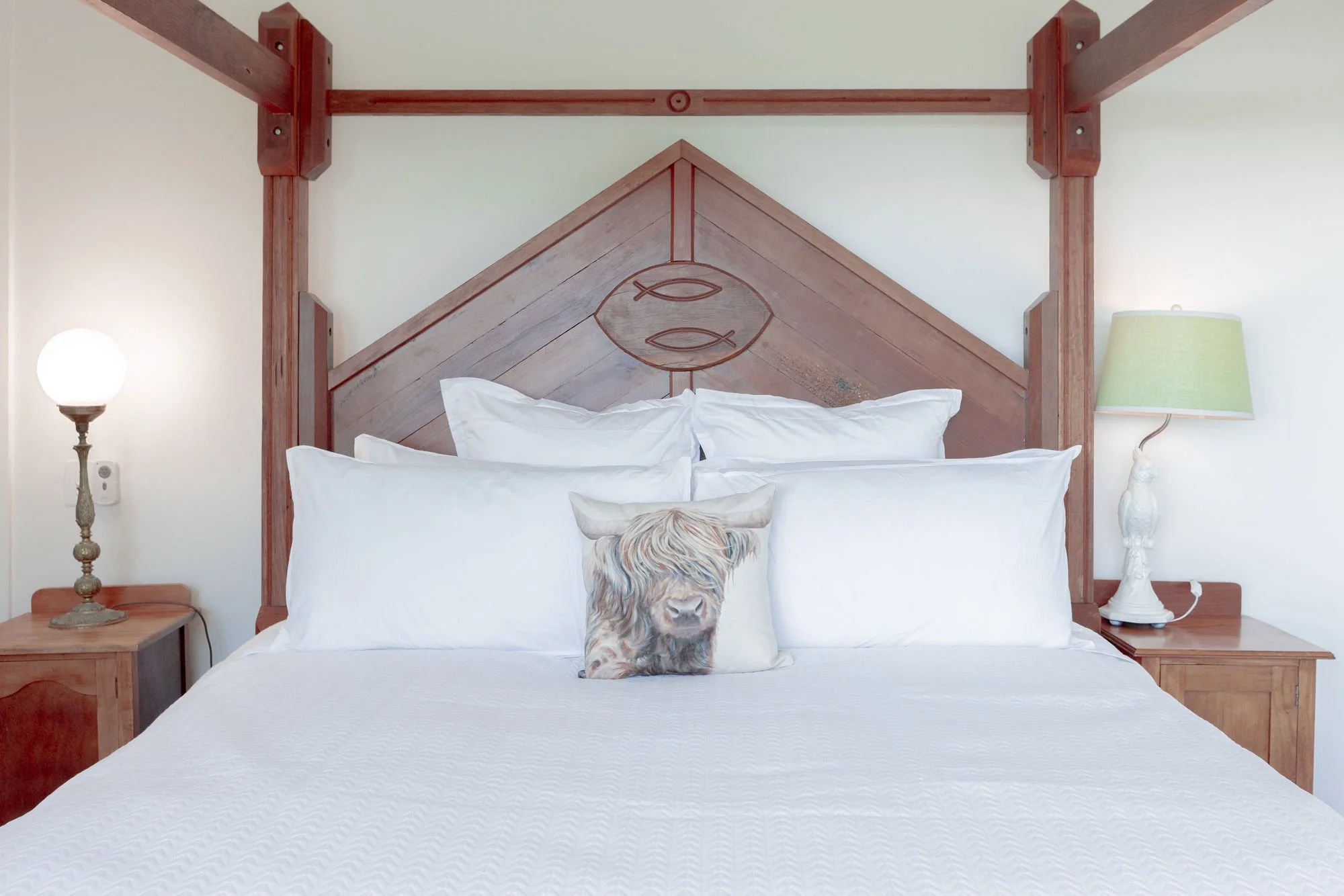 A bedroom with a wooden headboard featuring a fish symbol design, white bedding, and a decorative pillow with a dog image. There are two wooden nightstands with lamps on each side of the bed.