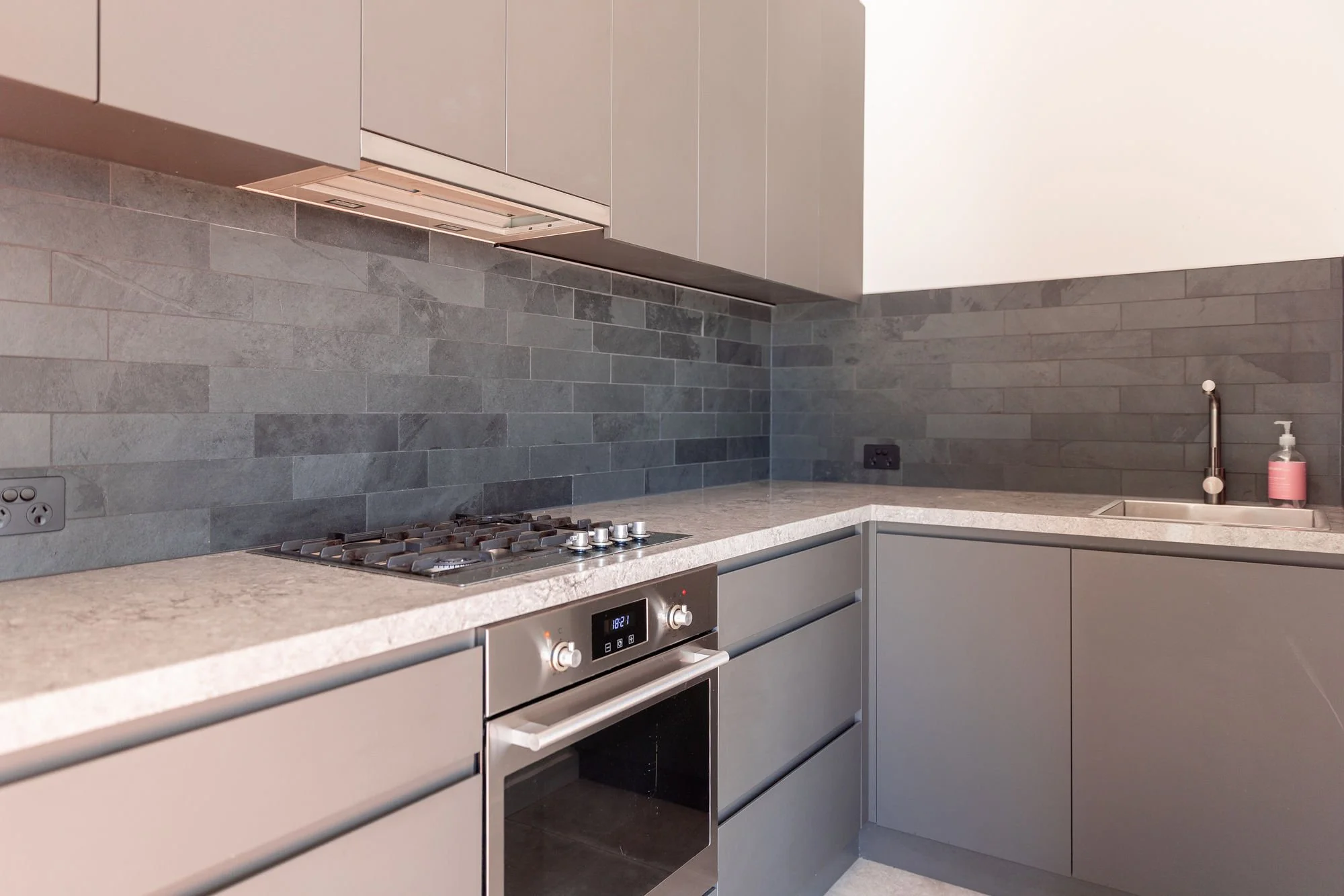 Modern kitchen with gray cabinets, gray stone backsplash, gray countertops, stove, oven, and a pink soap dispenser by the sink.